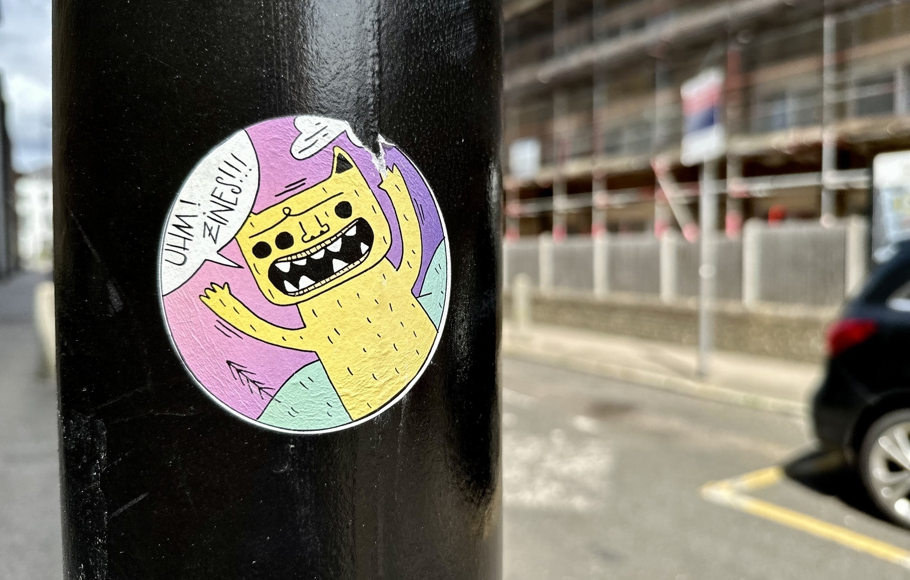 A round sticker on a black post. In the sticker, a yellow cartoon creature shouts UHM! ZINES!!!