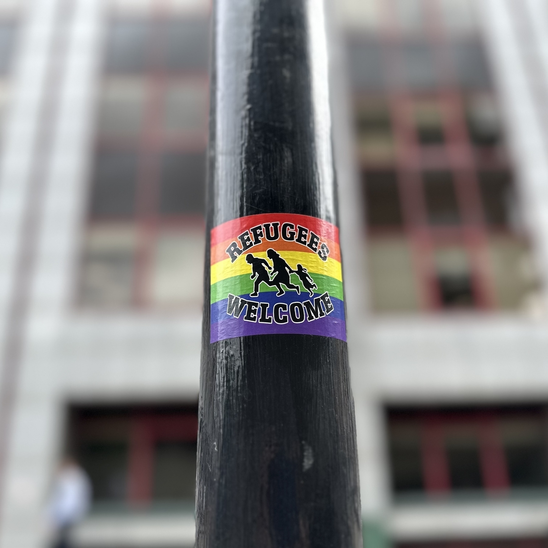 A sticker on a post that says Refugees Welcome over a rainbow flag background.