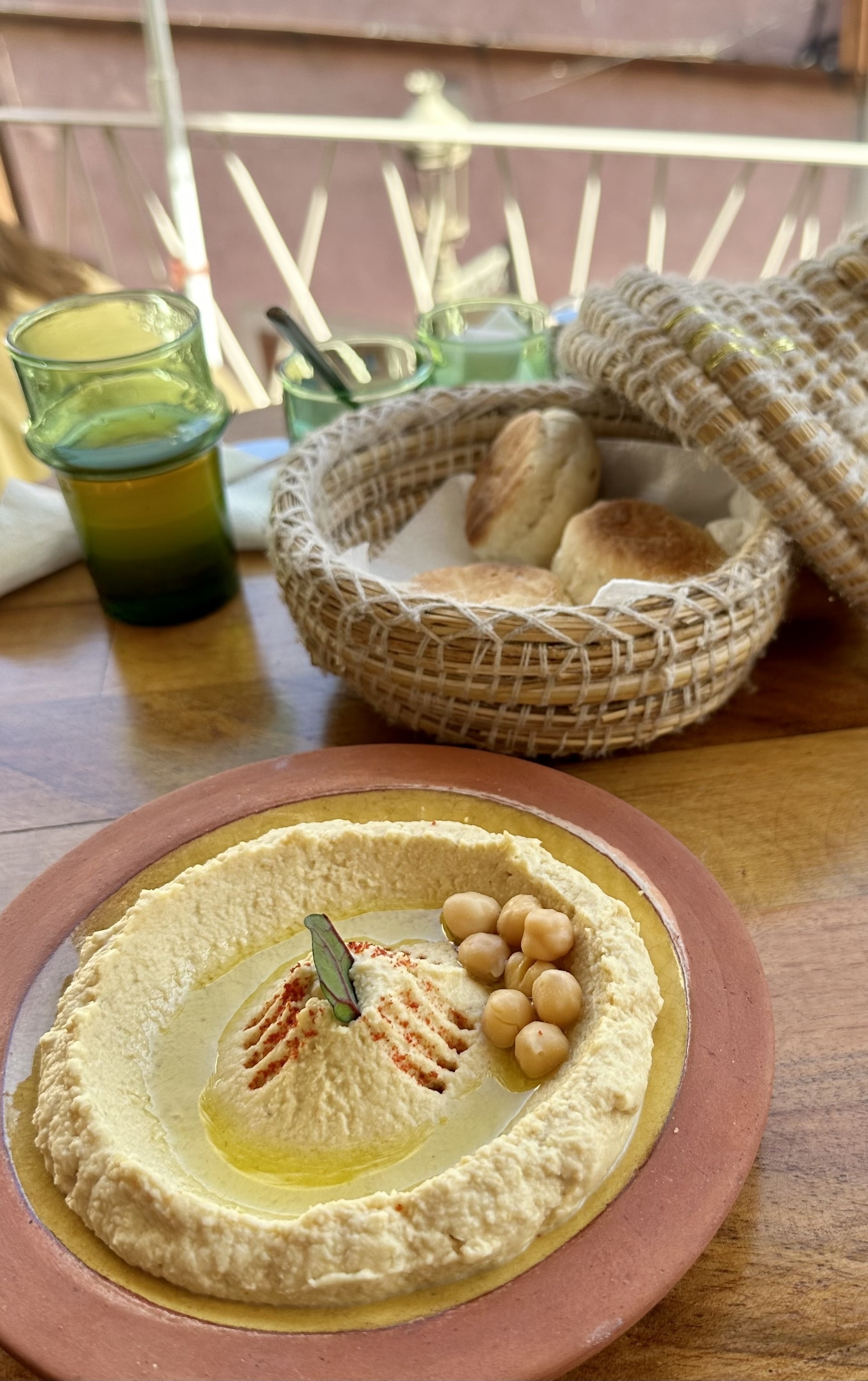 Hummus on a plate with some small bread rolls in a woven basket in the background, along with a glass of tea.
