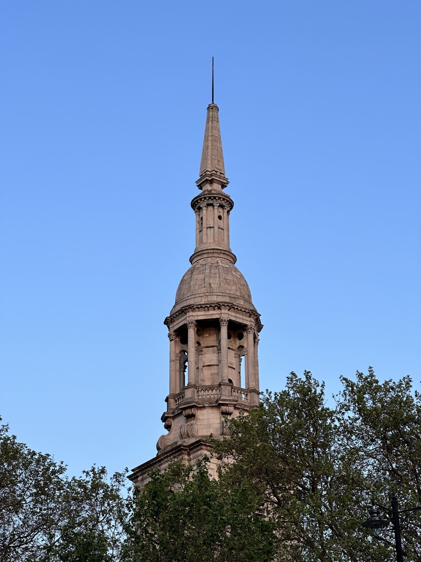 A stone church spire, lit with the warm light of sunset. Some trees are in the foreground and the background is a clear blue sky.