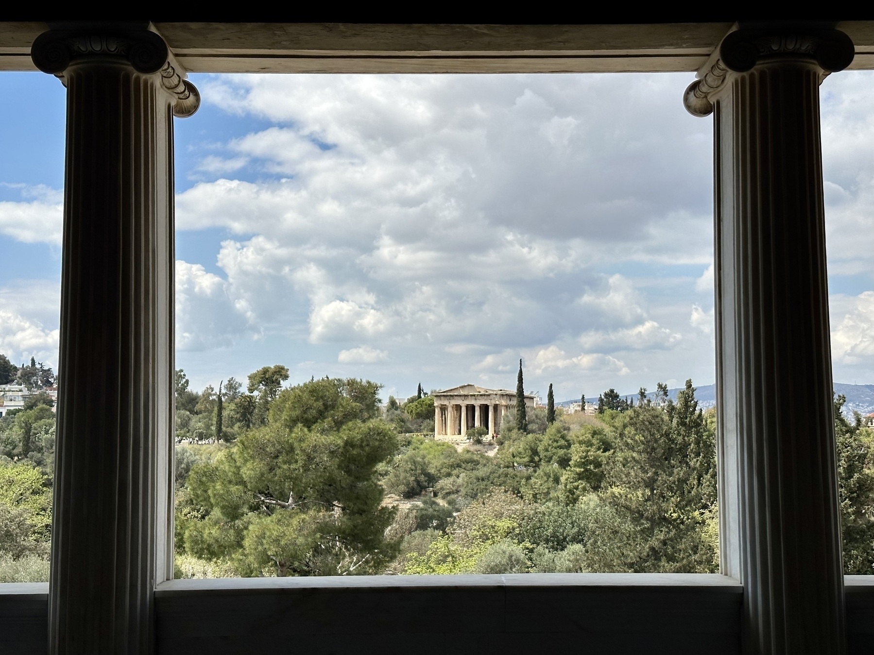 The temple of Hephaestus, viewed from the Stoa of Attalos, framed with an Ionic column on either side. The temple is in the distance on a hill, with trees in the foreground. 
