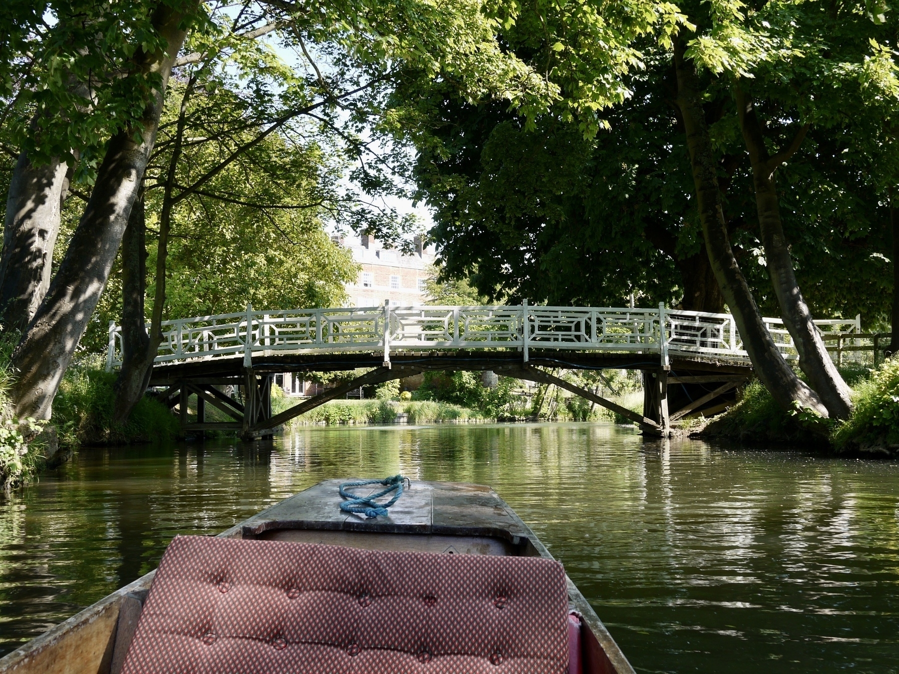 A punt on a tree-lined stretch of the River Cherwell in Oxford. A wooden bridge with white fencing crosses the river ahead of the punt.