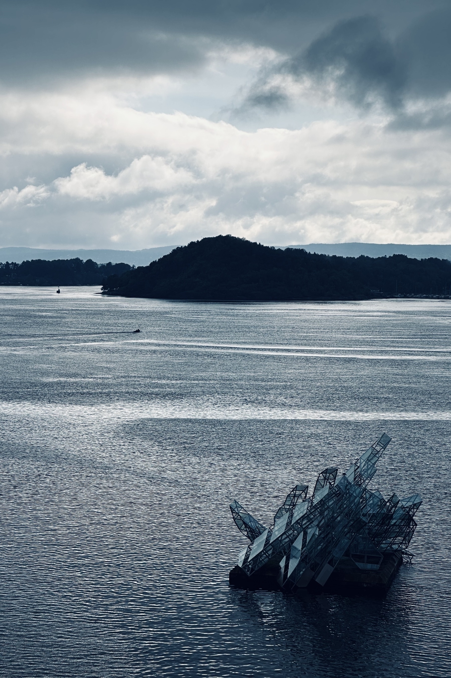 A sculpture made of shards of glass in Oslo harbour, with hills and a cloudy sky in the background.