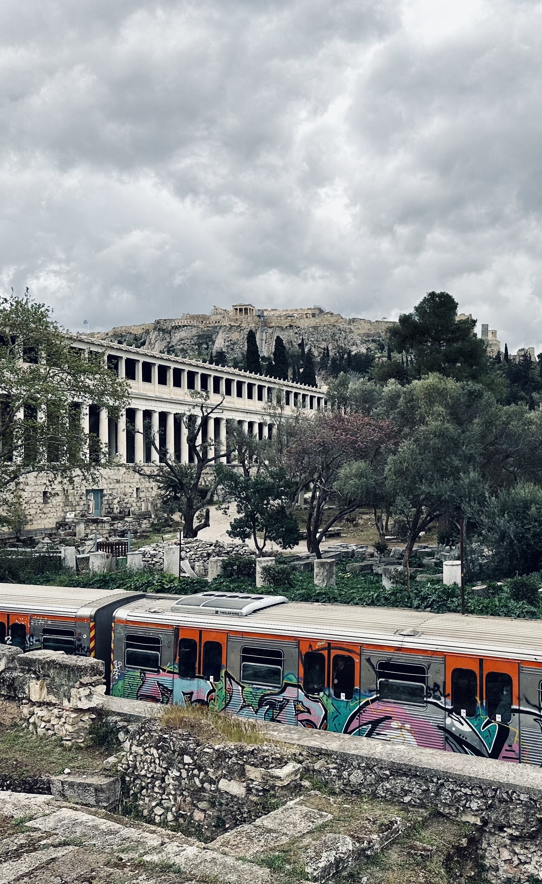 A metro train passing in front of the Stoa of Attalos in the Agora of Athens. In the distance is the Acropolis, with grey clouds above.