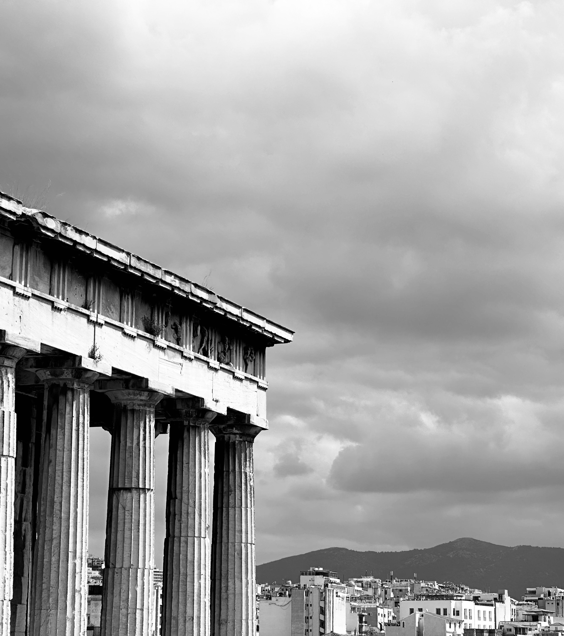 A Greek temple in the foreground, with modern buildings in the background, and hills in the distance. All under a cloudy sky.