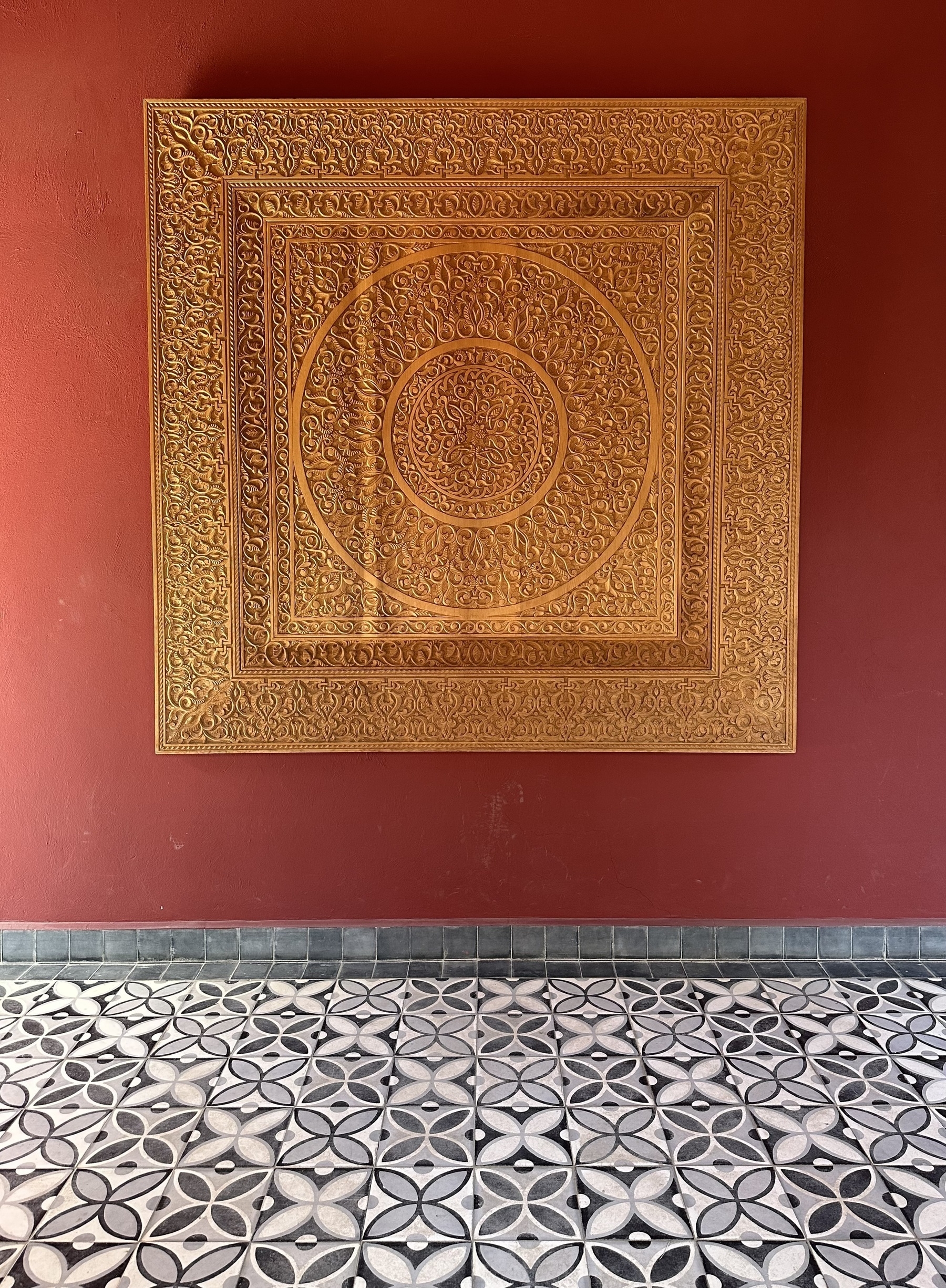 An intricately carved square wooden panel on a red wall, with black and white patterned tiles on the floor.