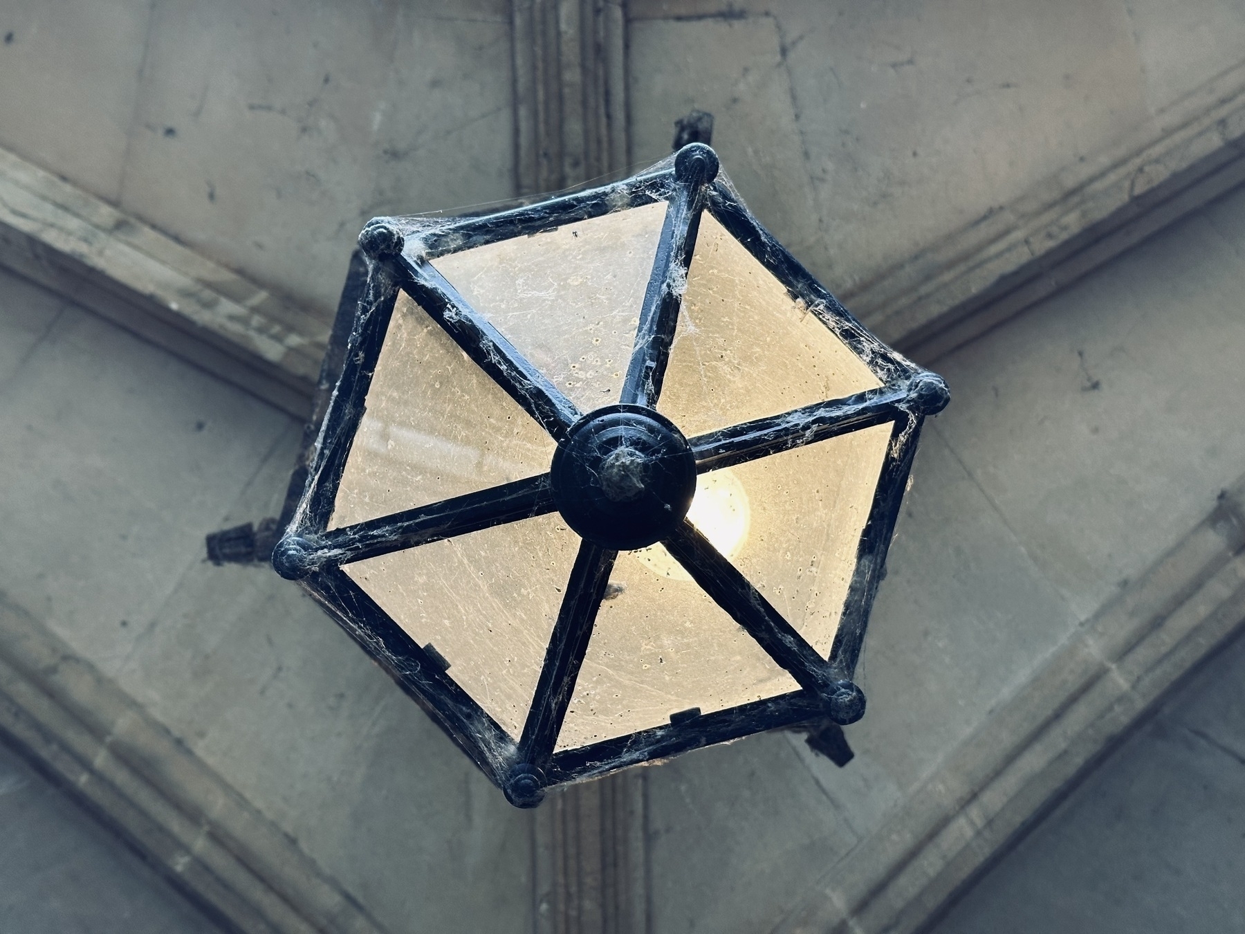 Looking directly up at a hexagonal metal framed lamp. The lamp has cobwebs around the edges, and is hanging from a vaulted stone ceiling.