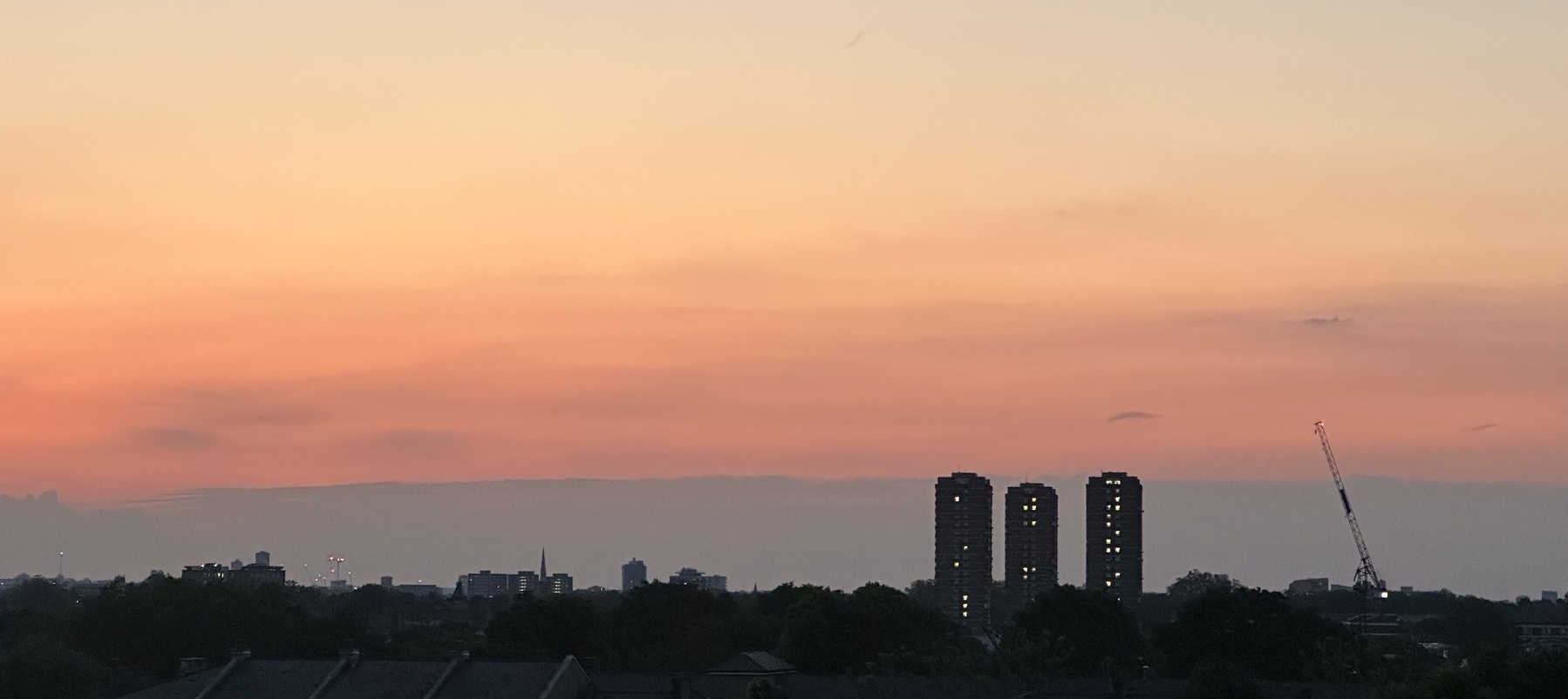 A photo of the sky at sunset. Darkish clouds are along the horizon, and above them a gradient changing from red to yellow in muted colours. Three tower blocks and a crane reach in to the sky on the right of the horizon.