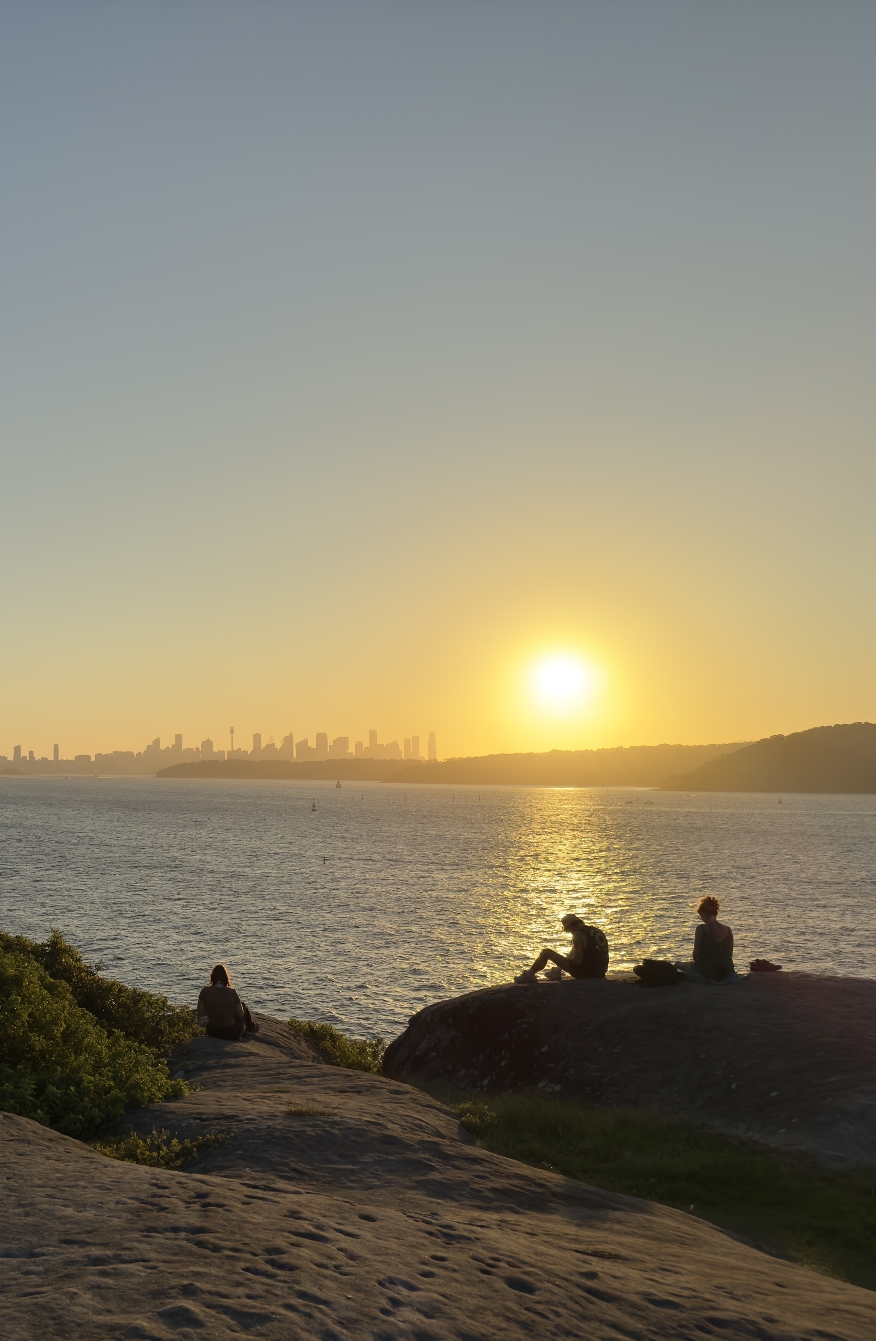 People sat on rocks watching the sun set over the Sydney skyline and water.