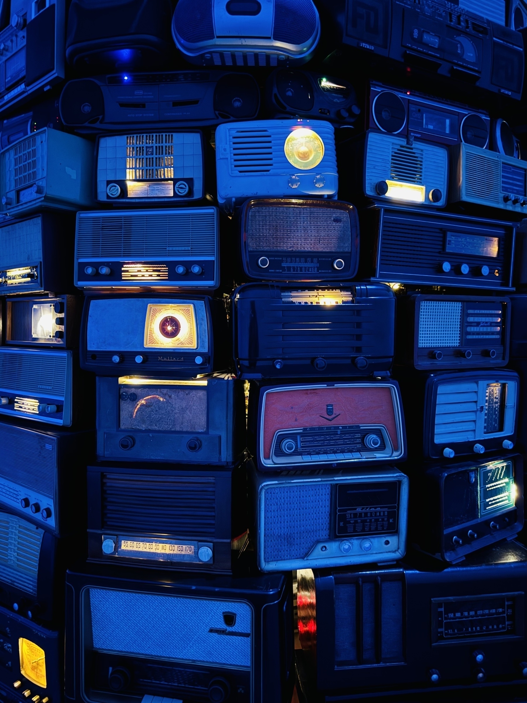 A collection of radios and tape decks stacked closely together and bathed in blue light.