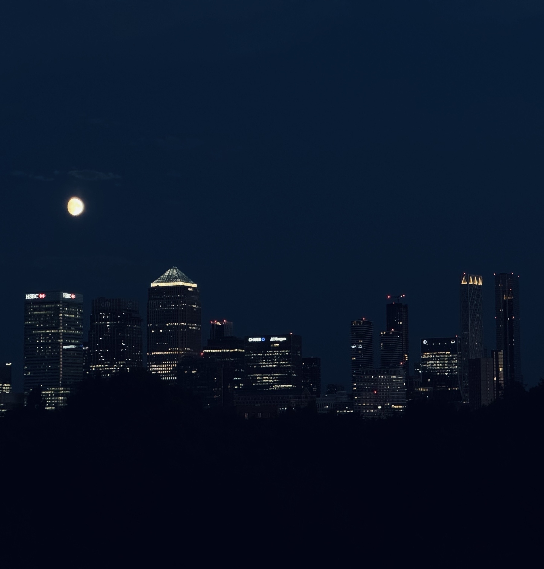 A bright moon in a dark blue sky above the skyscrapers of Canary Wharf.