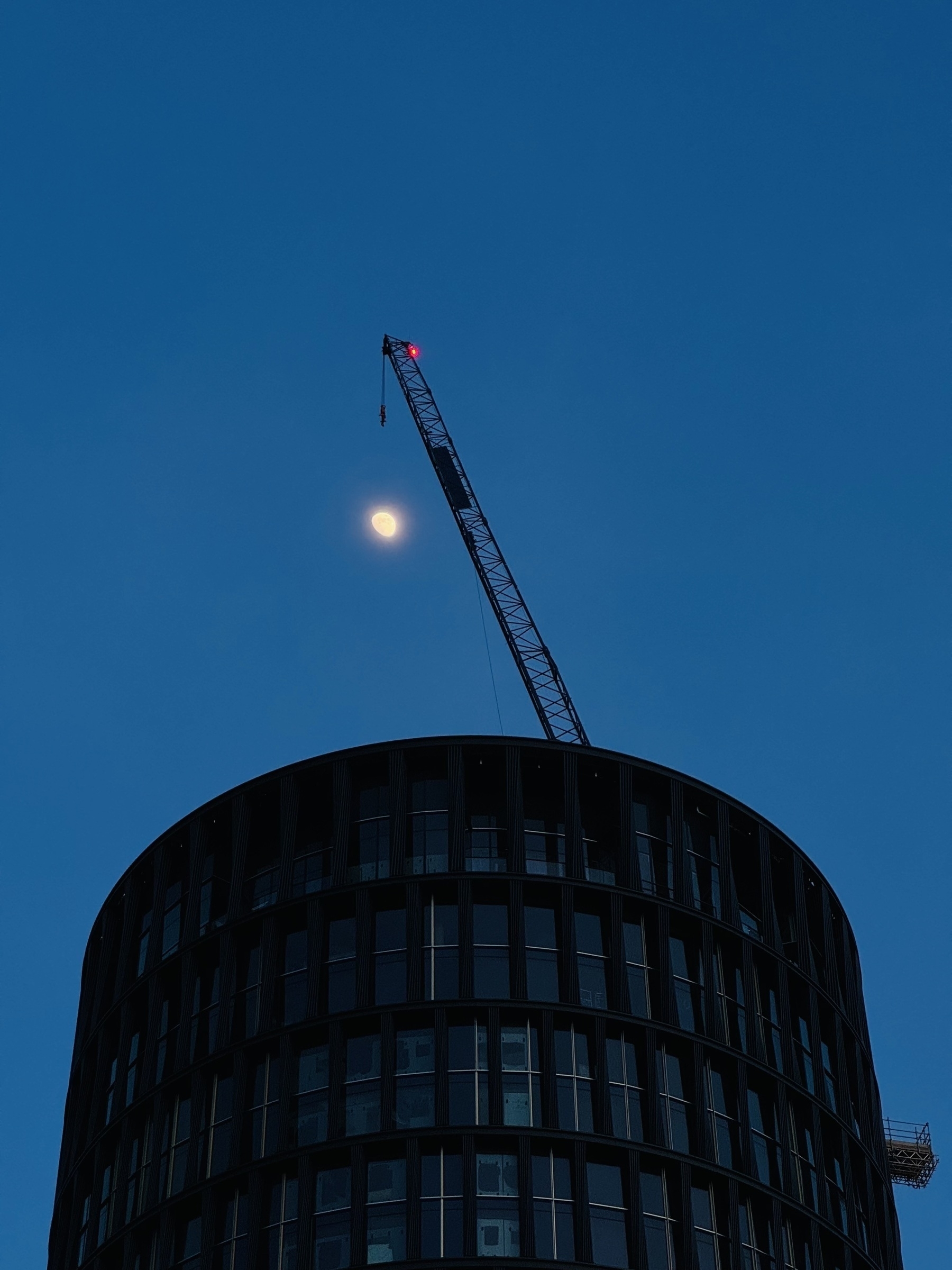 A crane poking out above a cylindrical building. The moon is hanging below the crane, making it look like the crane is fishing for the moon.