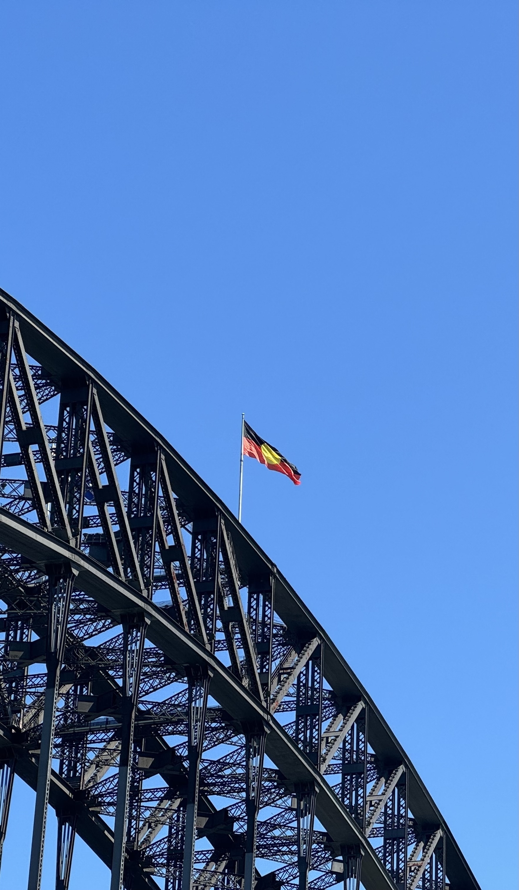 The Australian Aboriginal Flag (bottom half red, top half black, with a yellow circle in the middle) flying from the top of the Sydney harbour bridge. The sky is cloudless and blue.