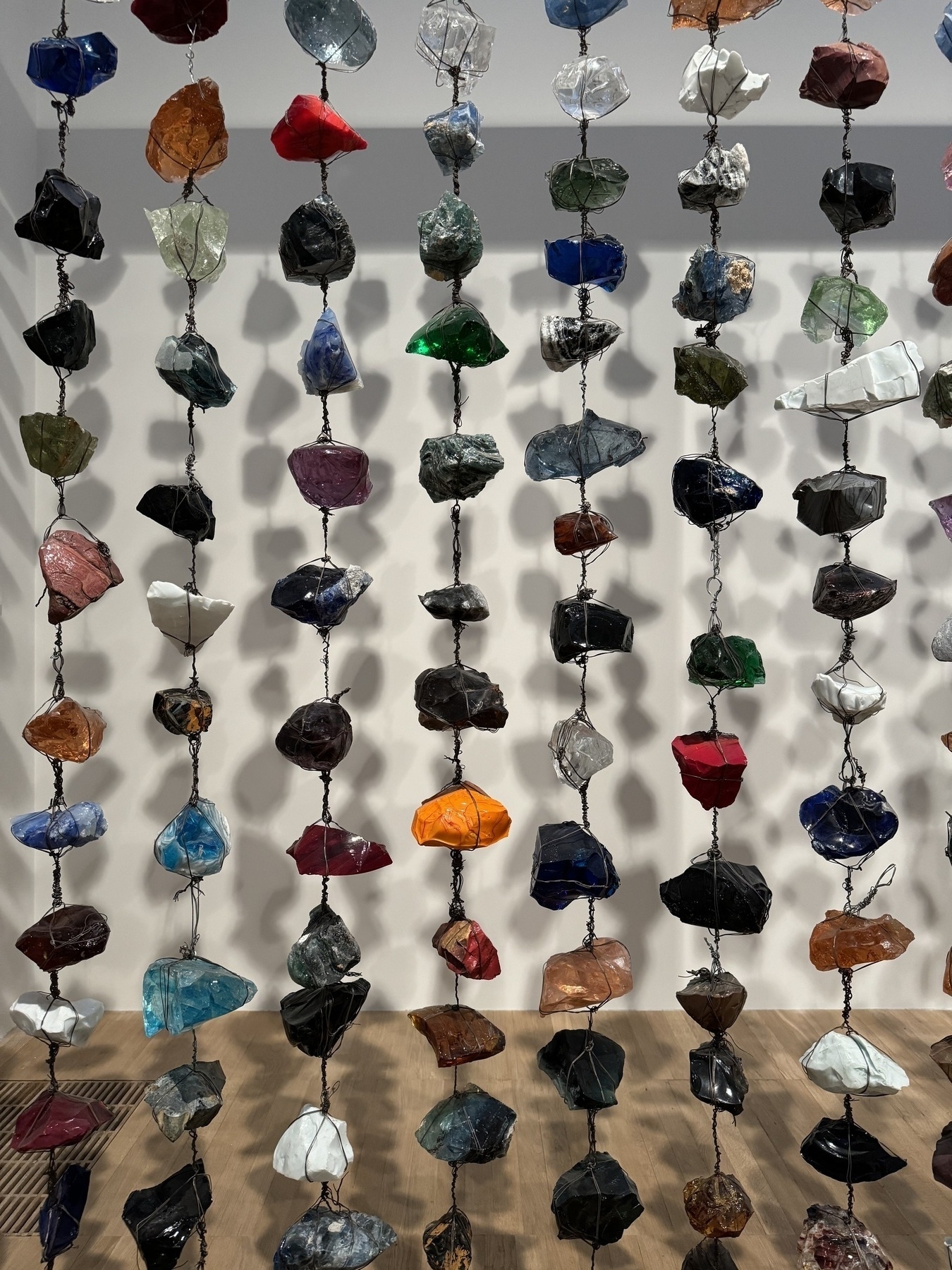 Rocks of varying colour suspended on a series of wires, casting shadows on to a white wall behind.