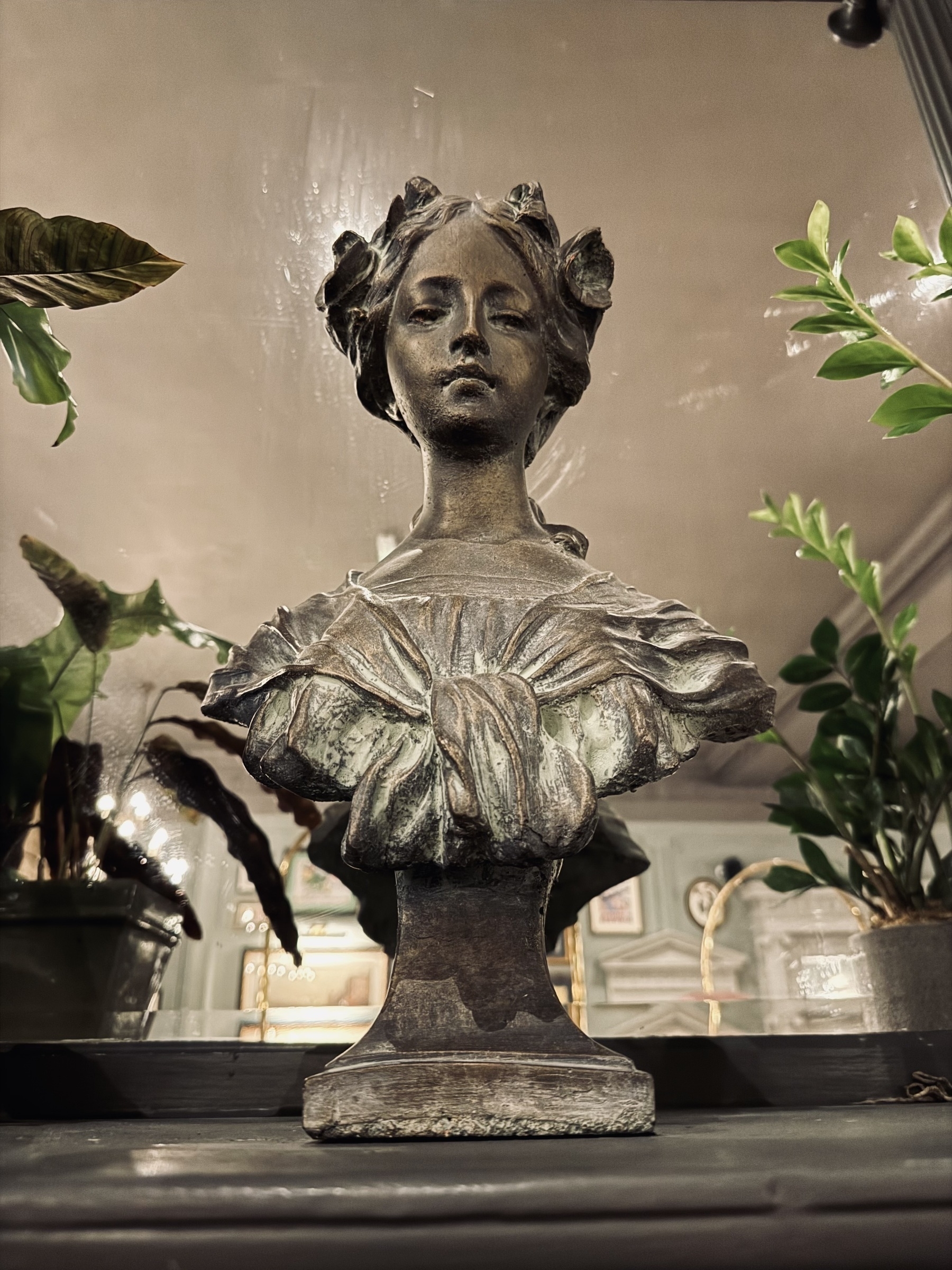 A bust sitting on a mantelpiece in front of a mirror. It’s surrounded by plants and looking down at the camera.