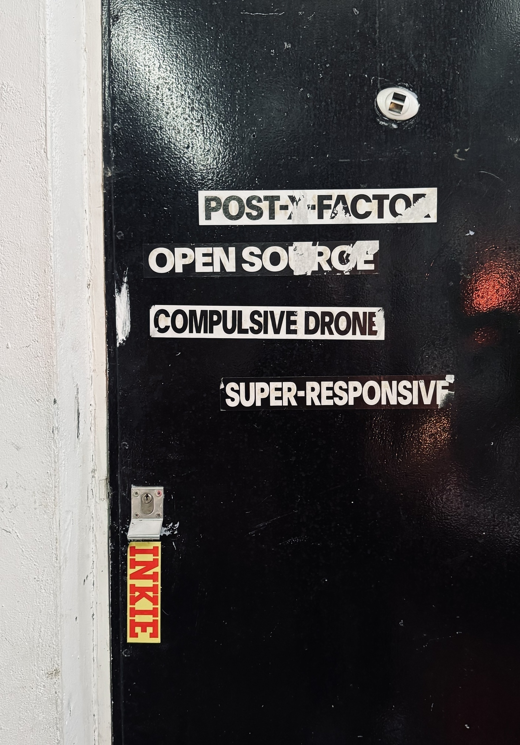 Stickers on a black door that say “post-x-factor”, “open source”, “compulsive drone”, and “super-responsive”.