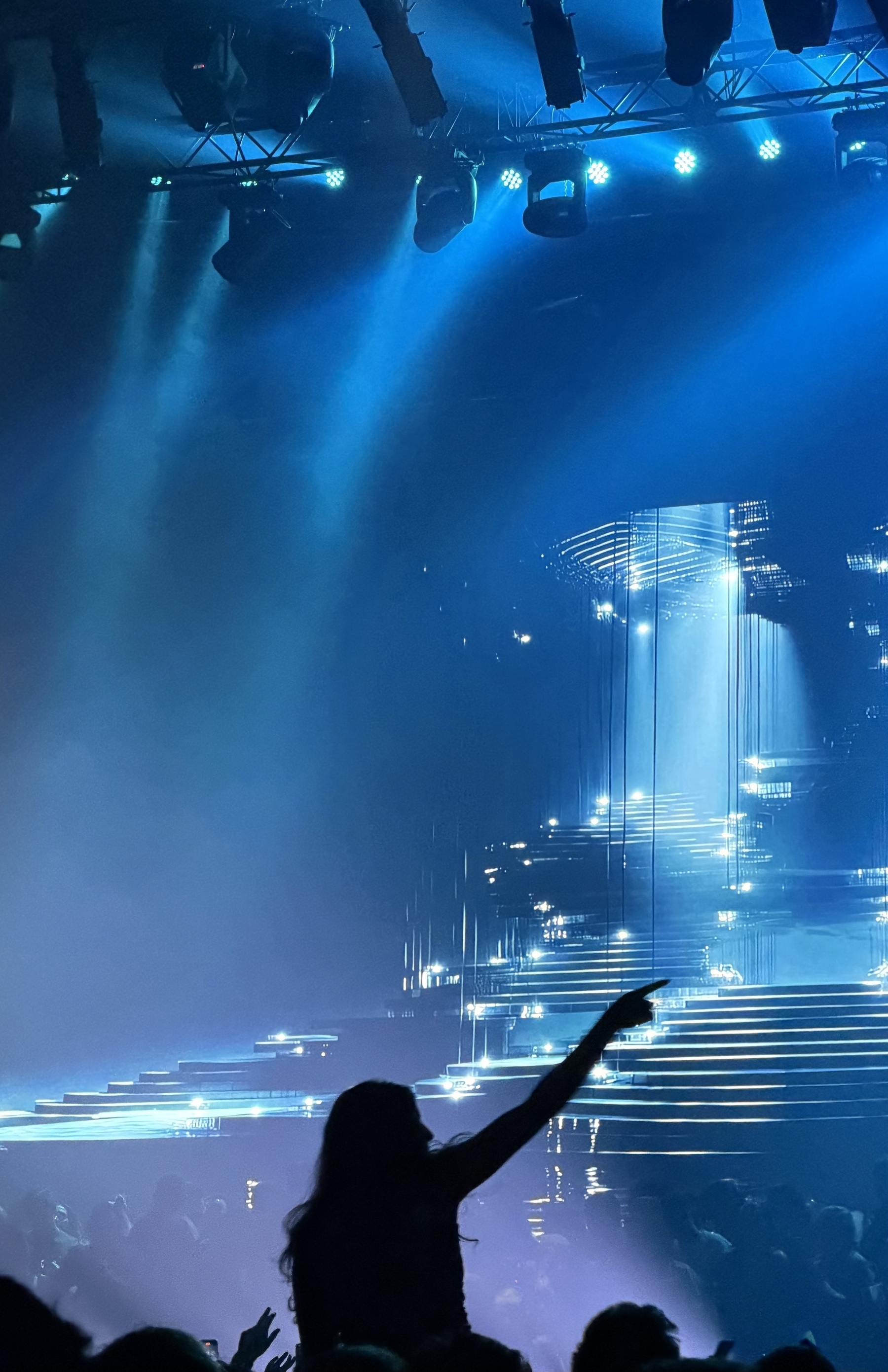 A huge video wall at a live music event, with lights above and a crowd at the bottom of the photo. One person in silhouette is raised above the others, with a hand raised in the air.