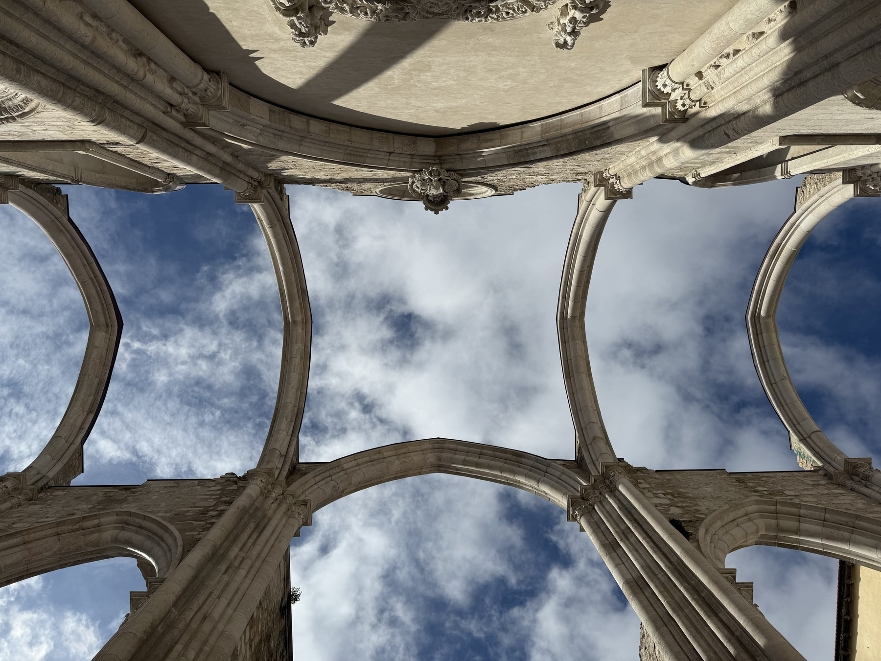 Looking up through the empty arches of Carmo Convent at white clouds in a blue sky.