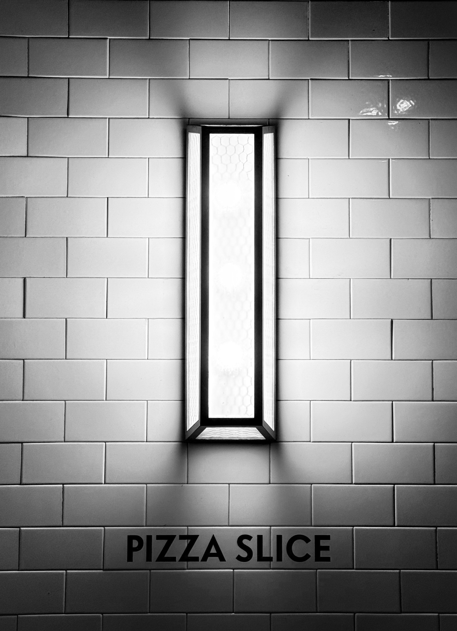 A lamp on a white tiled wall that says Pizza Slice below it.