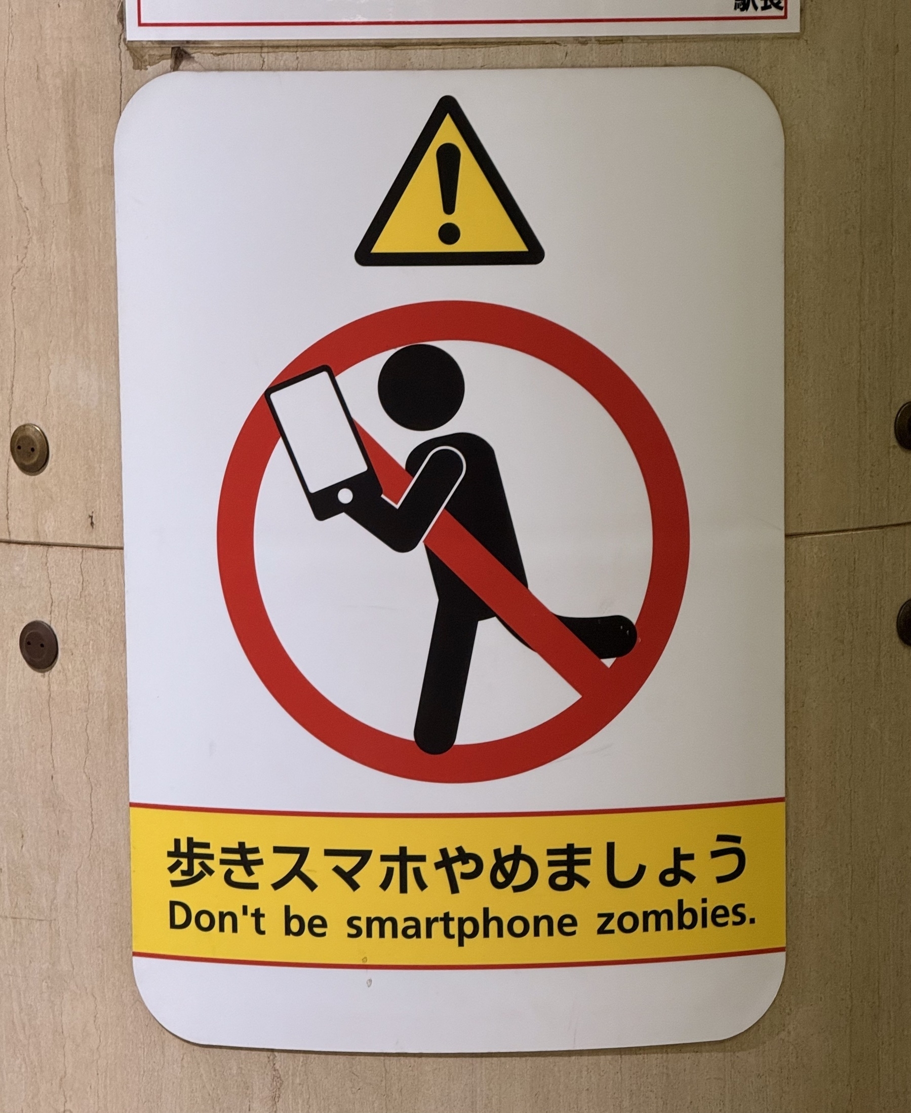 A sign on a column in a metro station in Tokyo. There’s a pictogram of a person walking while using a mobile phone in a red circle with a line through it. Underneath is a message that says “Don’t be smartphone zombies”.