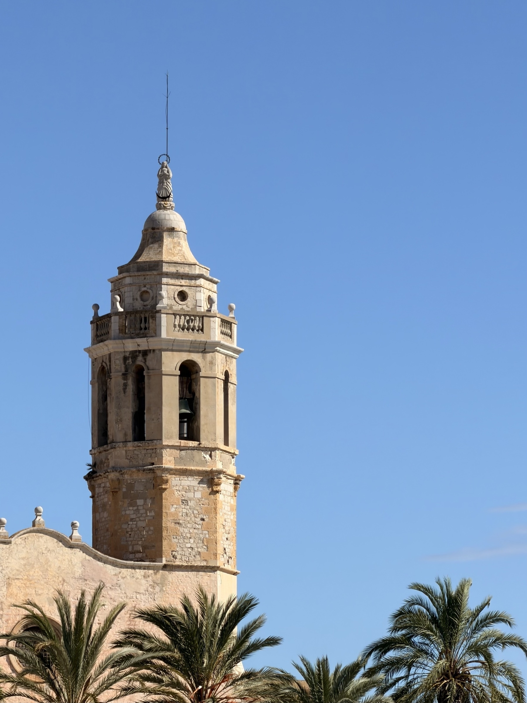 A stone church tower against a bright blue sky, with palm trees in the foreground.