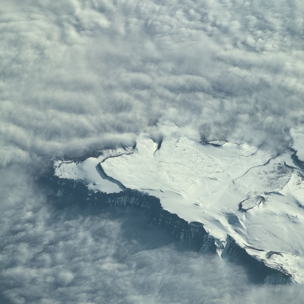 A snow covered plateau at the top of cliffs, rising out of the surrounding clouds.
