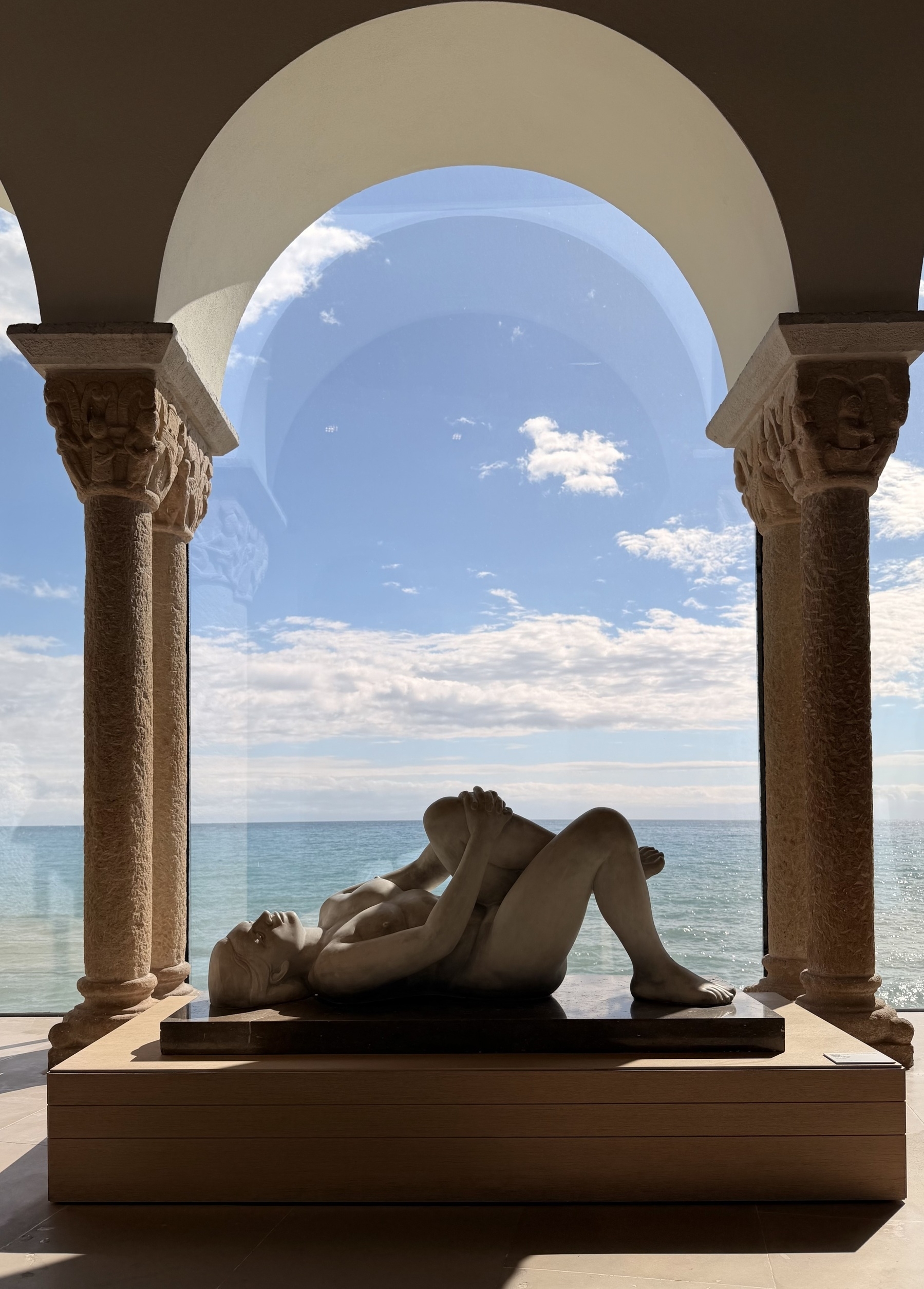 A statue of a naked woman lying on a plinth beneath an arch that has a view out over the sea.