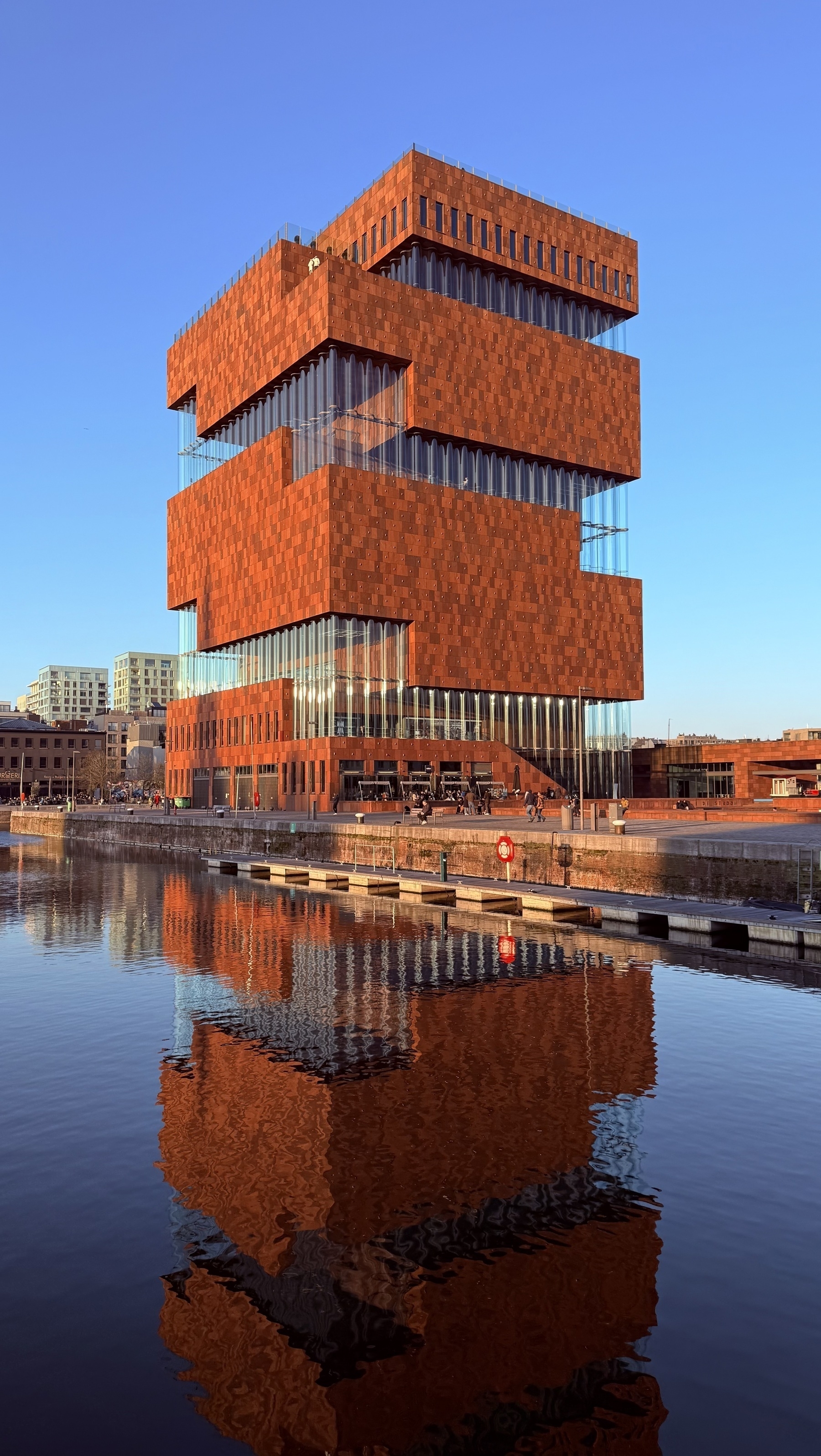 The stepped brick and glass facade of the Museum aan de Stroom in Antwerp, reflected in water.