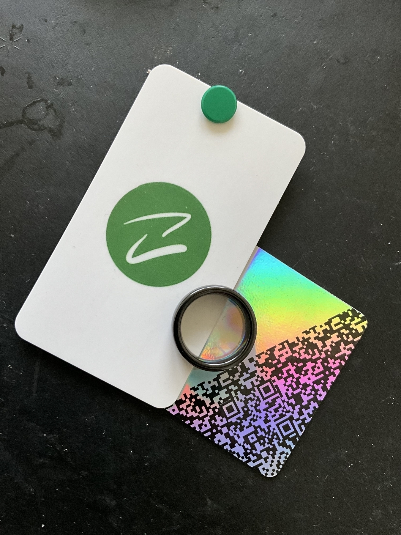 Black ring sitting on top of a white business card that shows Avanceé logo which sits on top of a business card that has a QR code and other symbols, looking like a QR code that are sitting on a holographic/rainbow colored business card. All items are sitting on top of a black background
