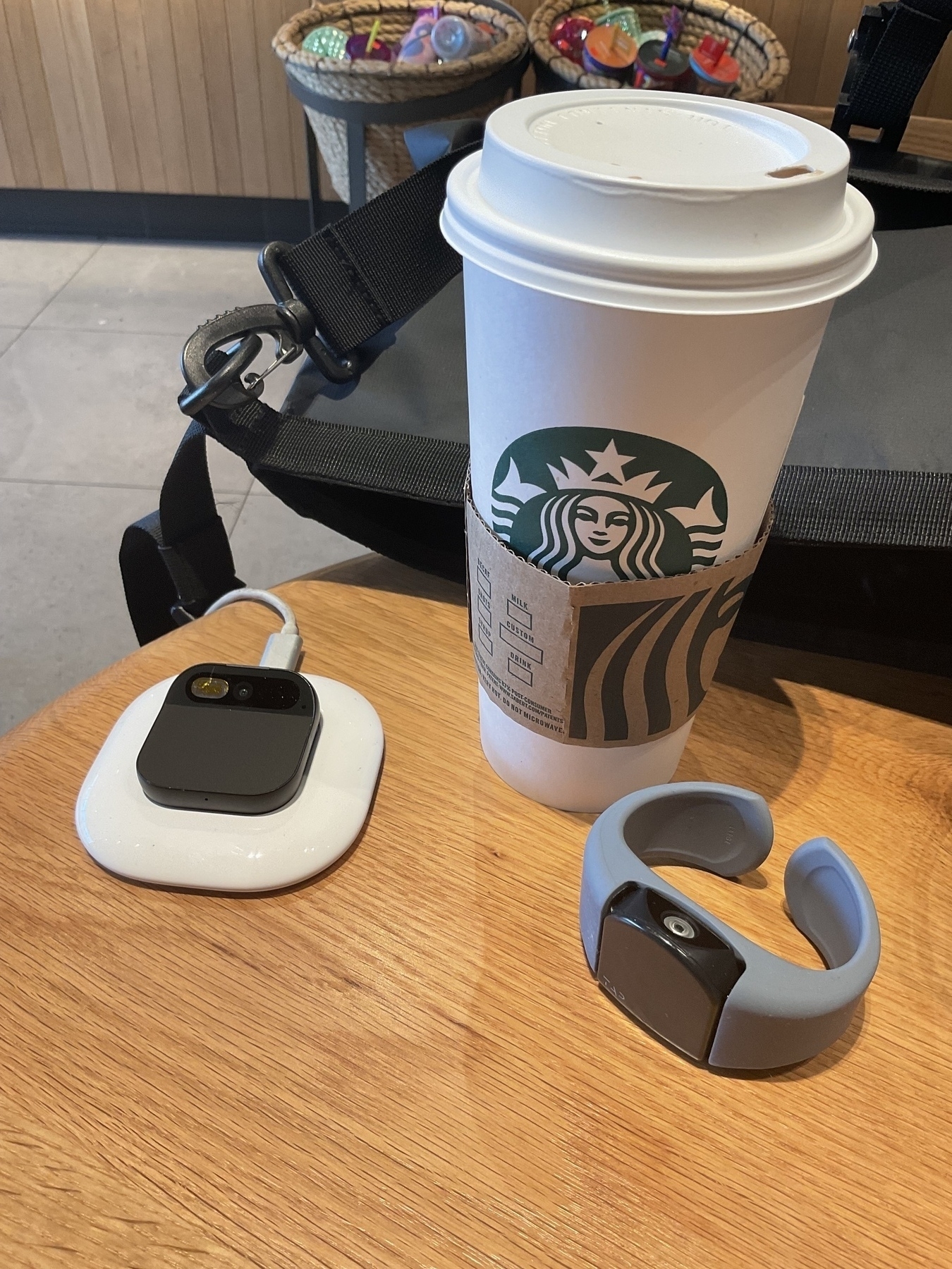 Humane AiPin and Tap wireless keyboard (right) in front of a Starbucks cup on a wooden table. Behind the cup is a bag, slightly open. Further back are baskets with product to be sold.