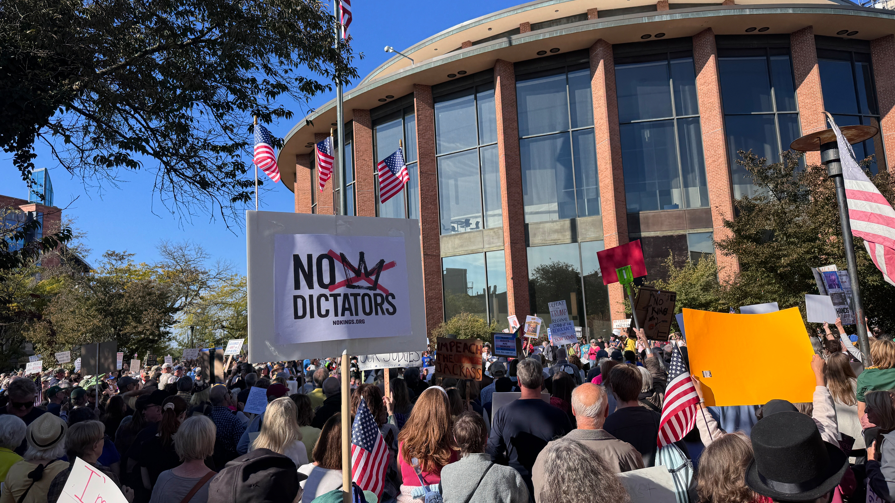A large crowd is gathered in front of a building, holding signs and American flags at a protest.