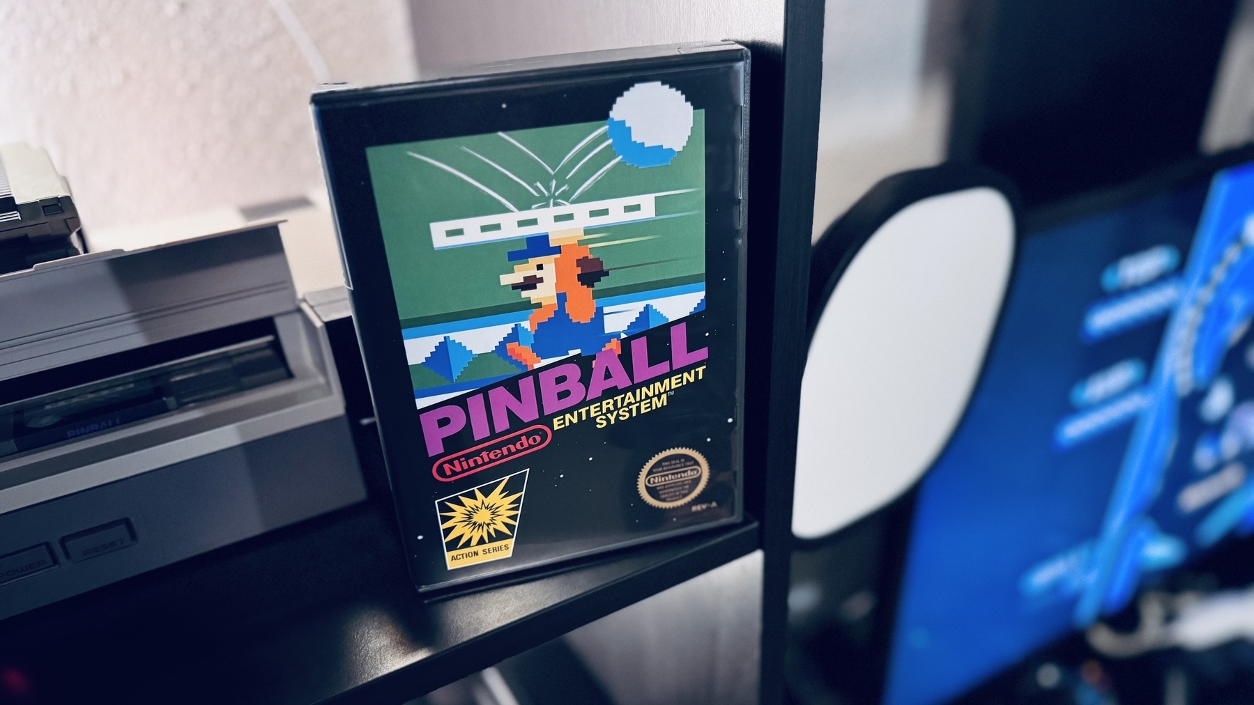 Auto-generated description: A boxed Nintendo Pinball game featuring a colorful 8-bit design is displayed on a shelf beside some gaming equipment.
