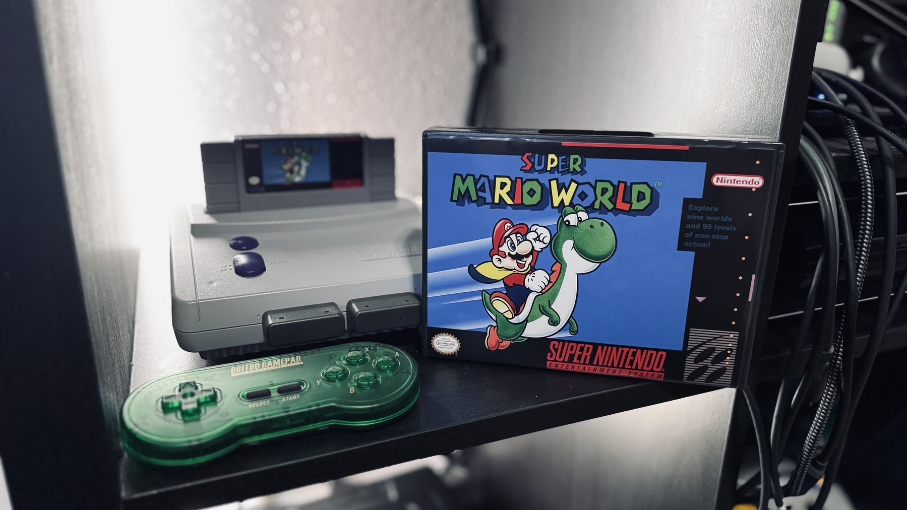 Auto-generated description: A Super Nintendo Entertainment System with a green controller and a boxed Super Mario World game is displayed on a shelf.