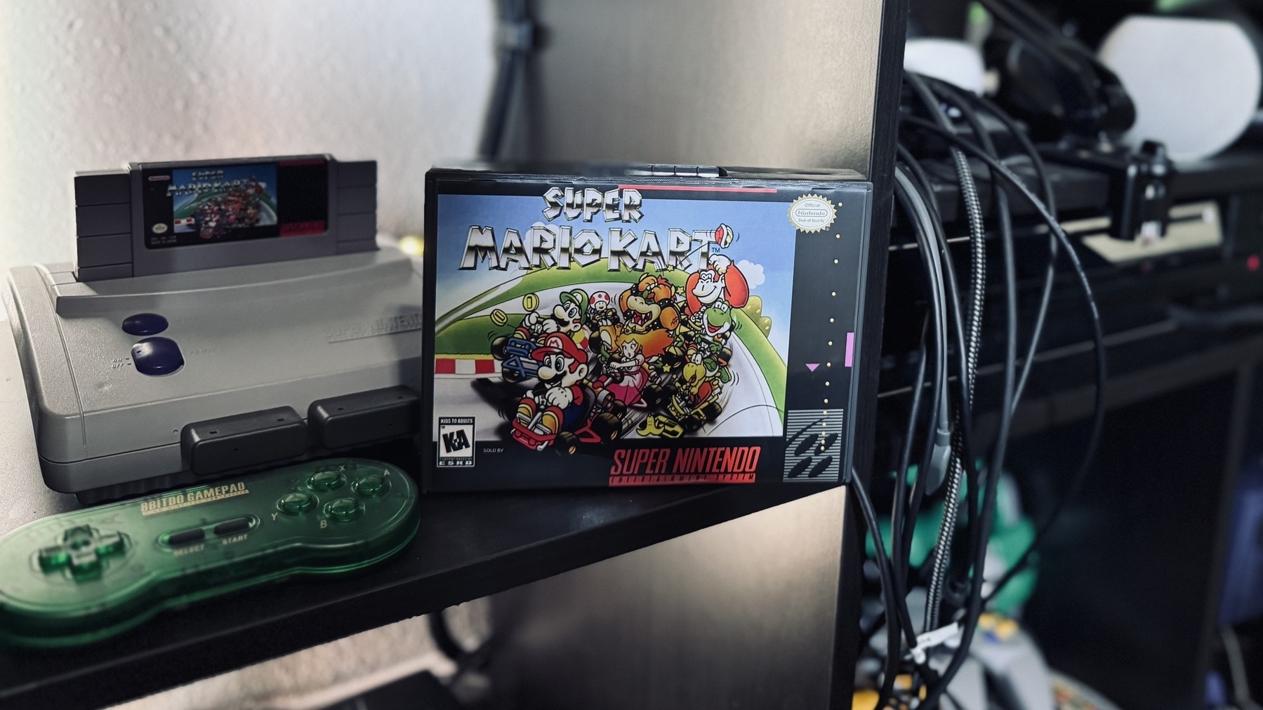 Auto-generated description: A Super Nintendo Entertainment System (SNES) with a green controller and a boxed Super Mario Kart game is displayed on a shelf.
