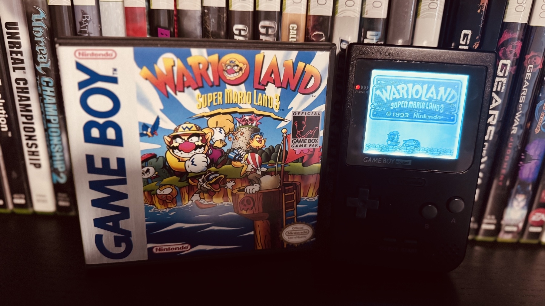 Auto-generated description: A Game Boy game case for Wario Land: Super Mario Land 3 is displayed next to a Game Boy showing the game’s title screen, with various video game cases in the background.