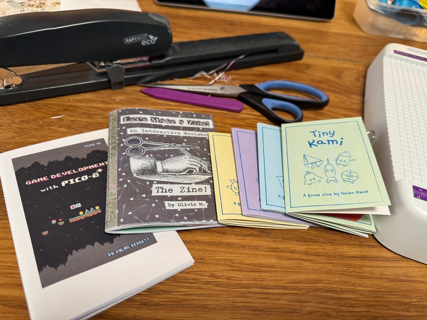 A collection of colorful zines and stationery items, including a stapler and scissors, are arranged on a wooden table.