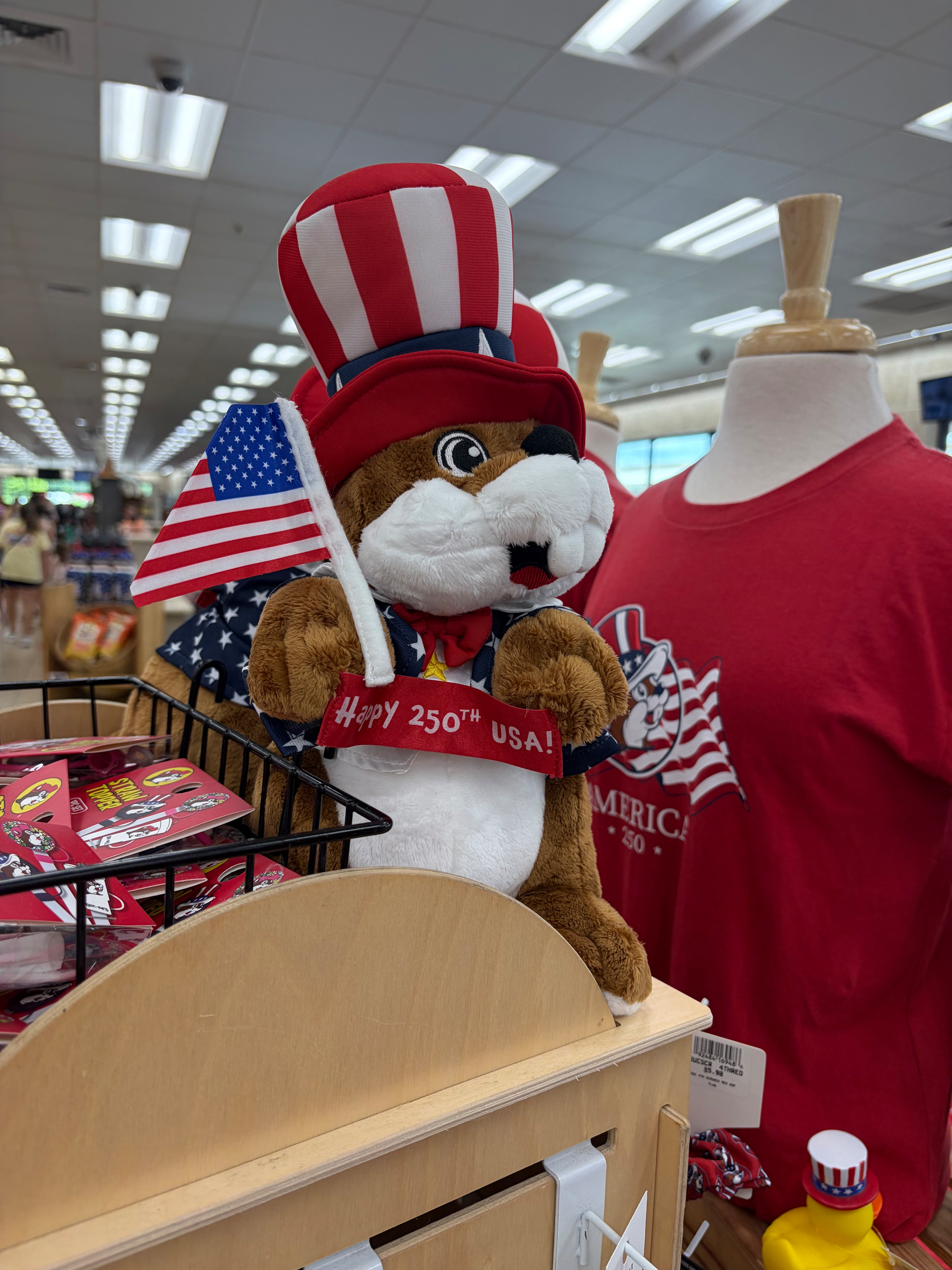 A stuffed animal wearing a patriotic hat and holding an American flag stands beside a mannequin dressed in a red T-shirt inside a store.
