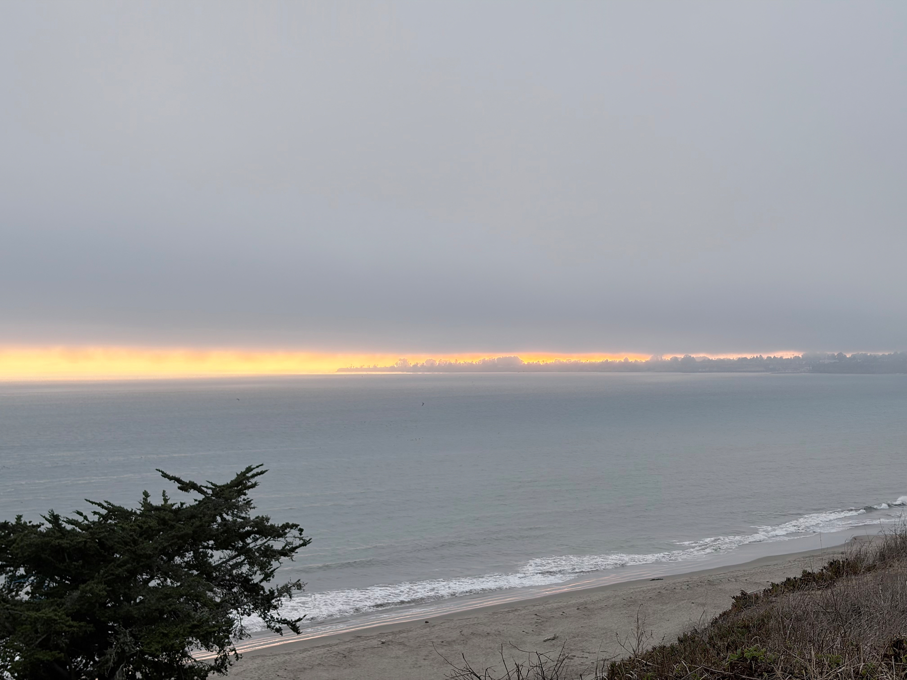 A calm ocean meets a sandy beach under a gray sky with a thin strip of golden light on the horizon.