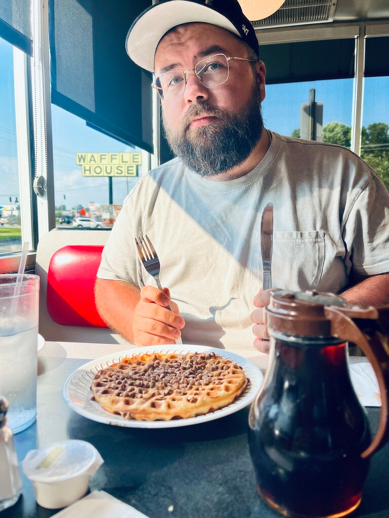 Shane Duffner sits at a table in a Waffle House, holding a fork and knife in front of a waffle topped with peanut butter chips.