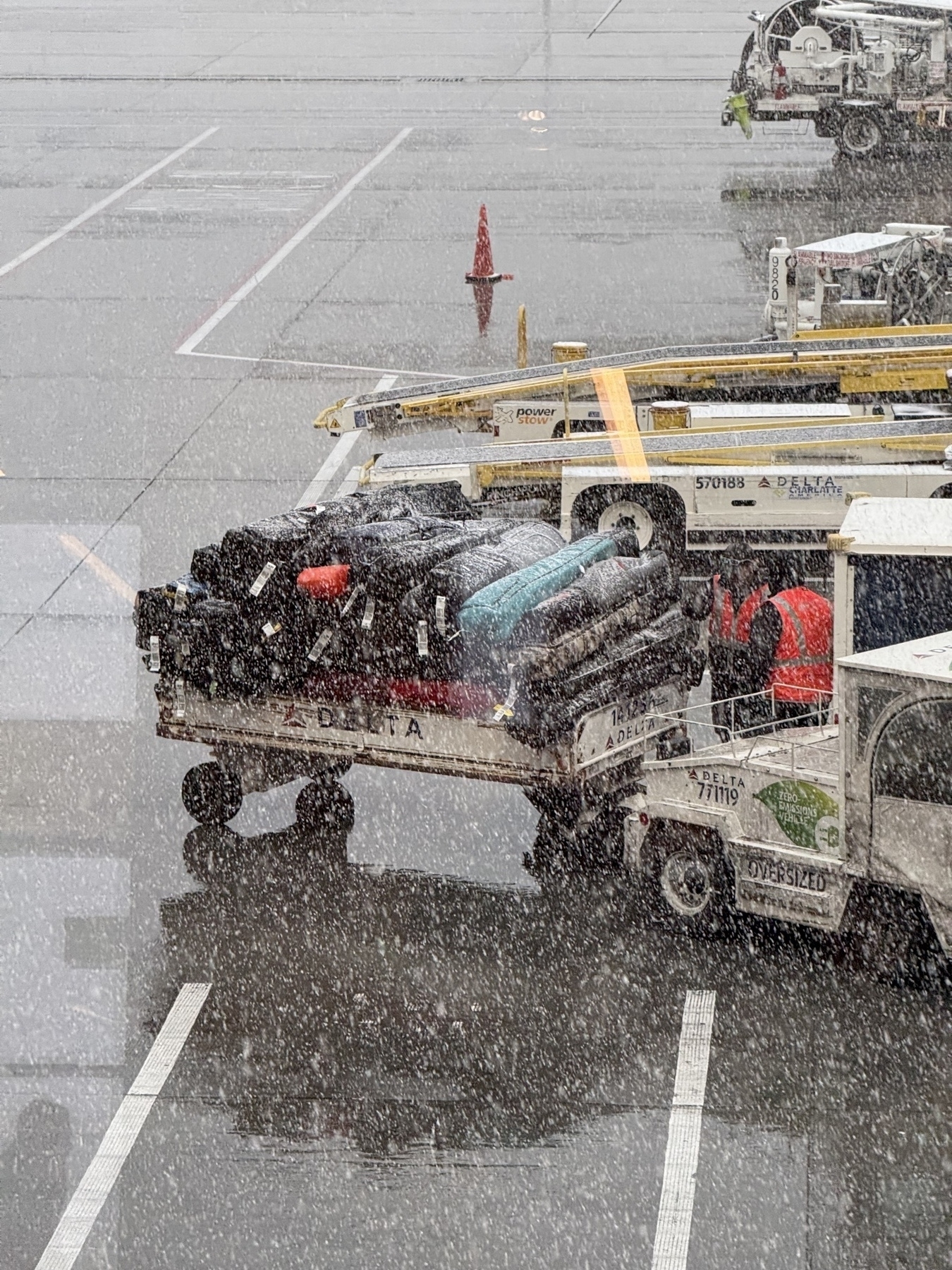 Picture of a luggage trailer on the tarmac near a plane. Trailer is loaded with dozens of ski bags. It’s snowing hard. 