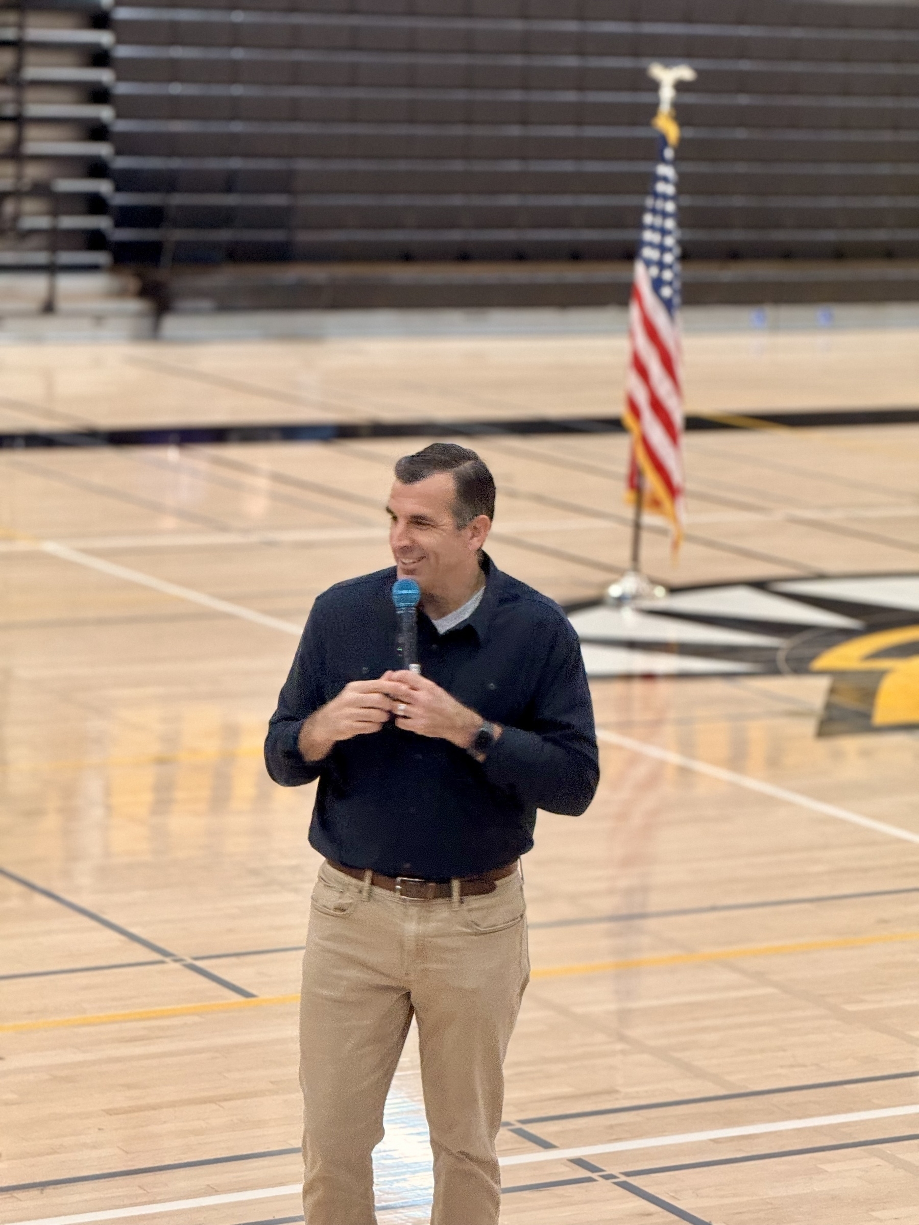 US Representative Sam Liccardo, holding a microphone, standing on a gym floor. US flag in the background. 