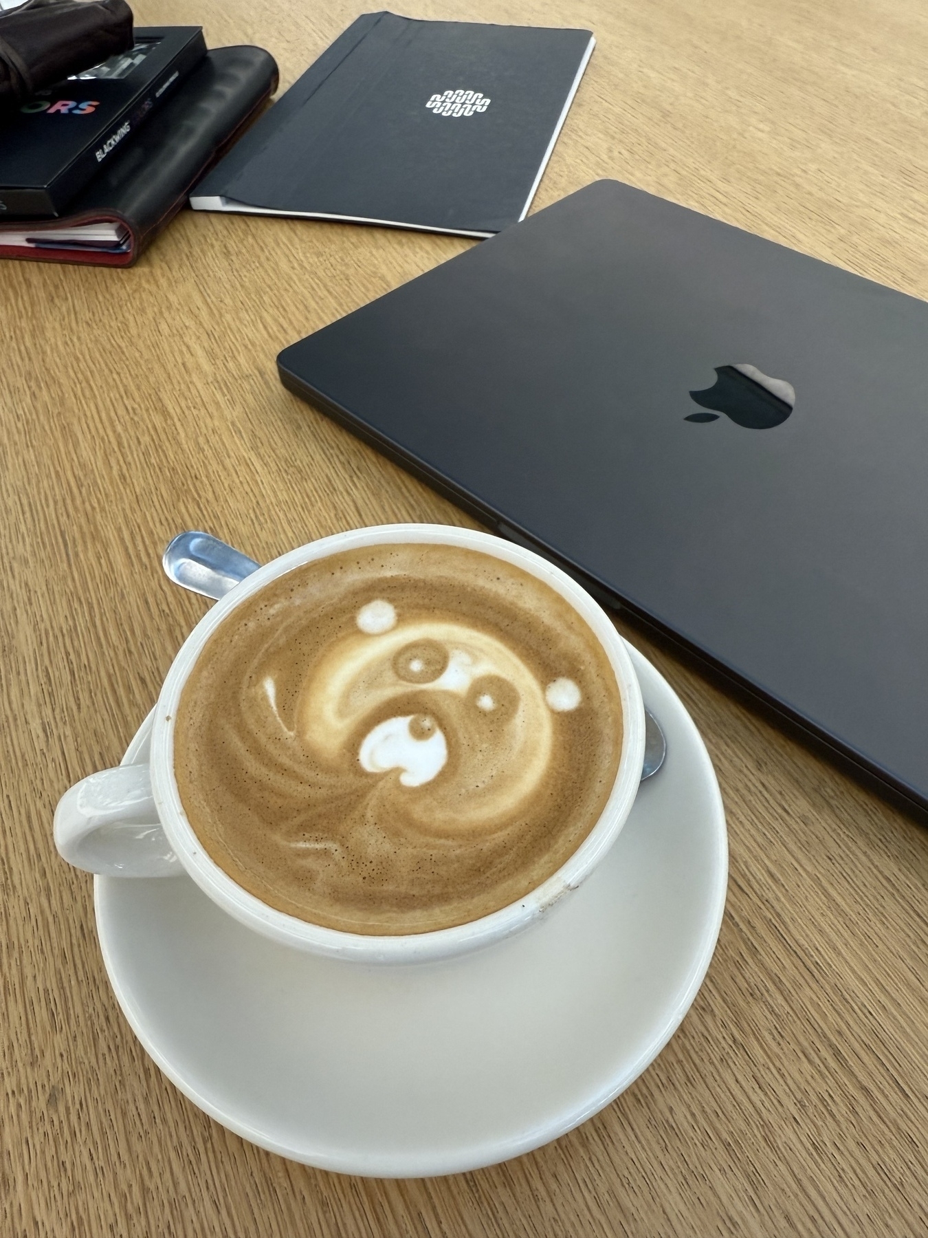 Cappuccino with bear-themed foam art. MacBook Pro, Sidekick notepad, planner, and stationery in the background. All on a very Apple maple table. 