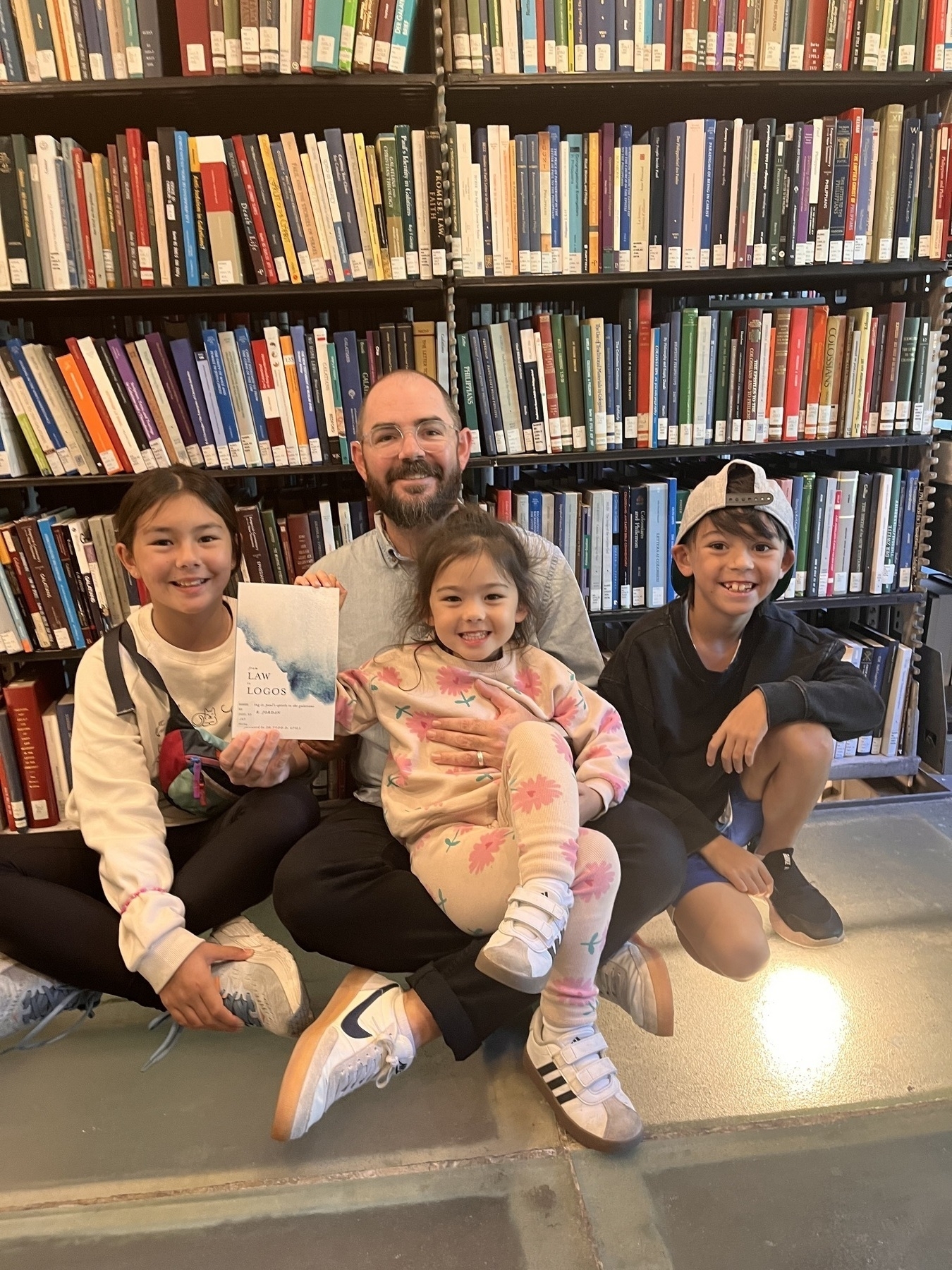 A man and three children are sitting together in a library, with the man holding a book.