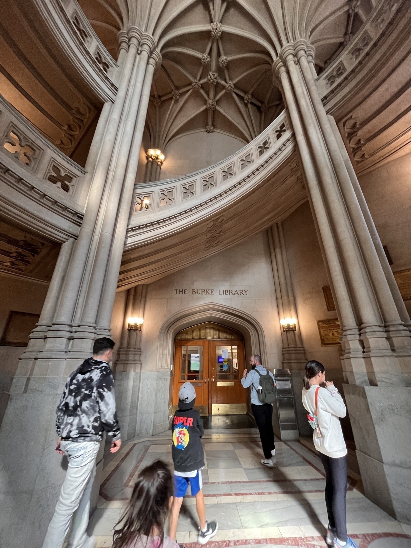 A group of people is approaching the entrance to a grand, ornate library with tall pillars and a vaulted ceiling.