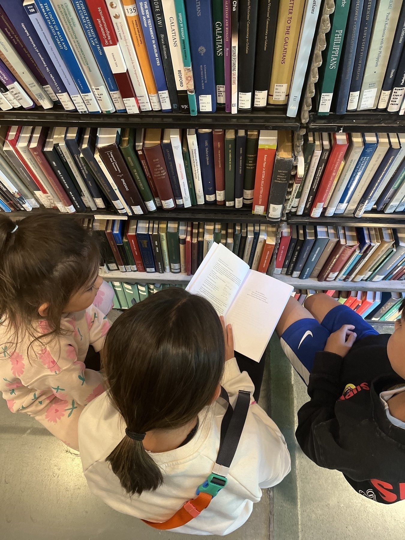 Three children are sitting together on the floor of a library, looking at a book in front of a bookshelf.