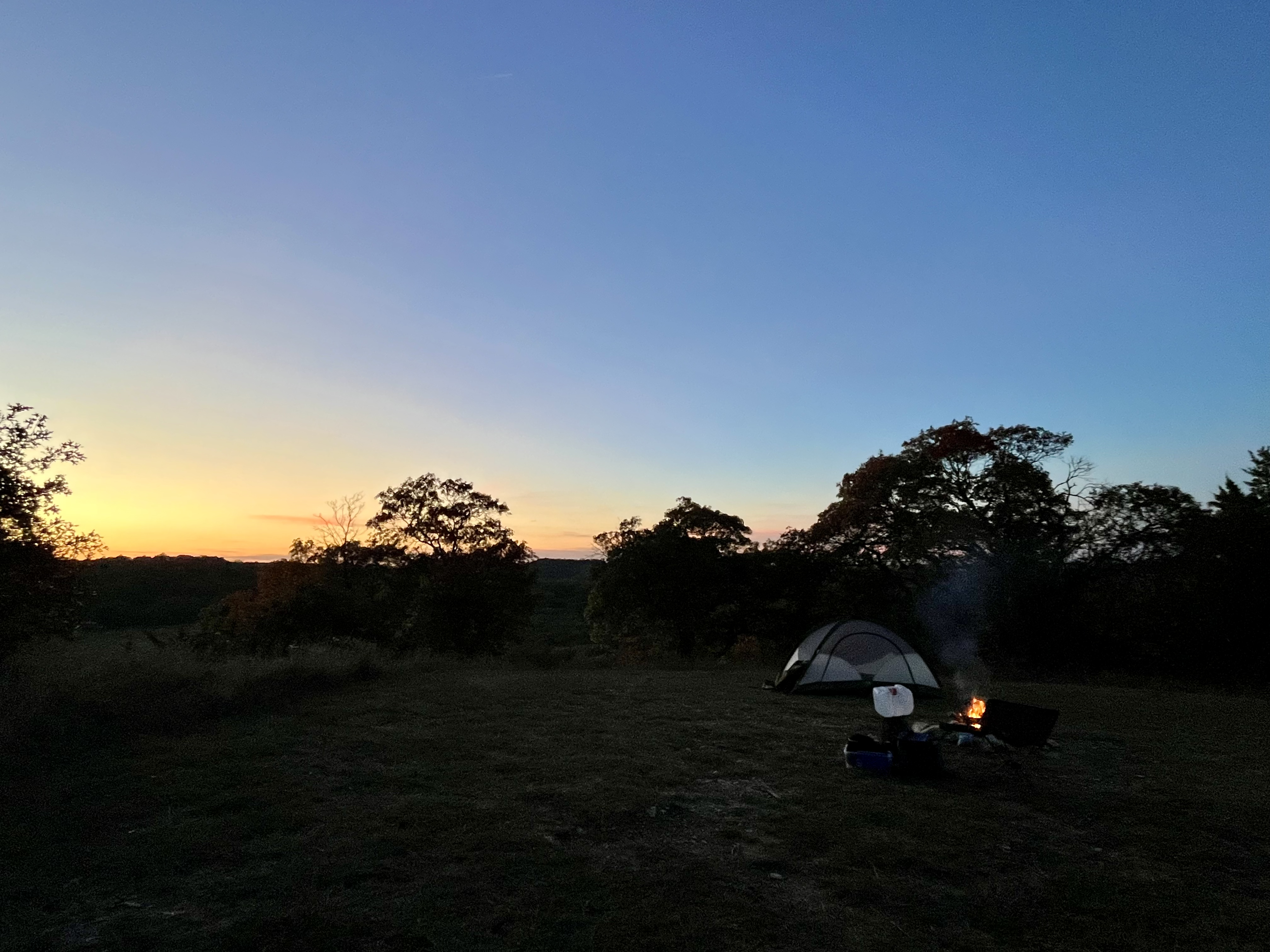 A peaceful campsite at dusk features a pitched tent and a campfire under a fading sky.