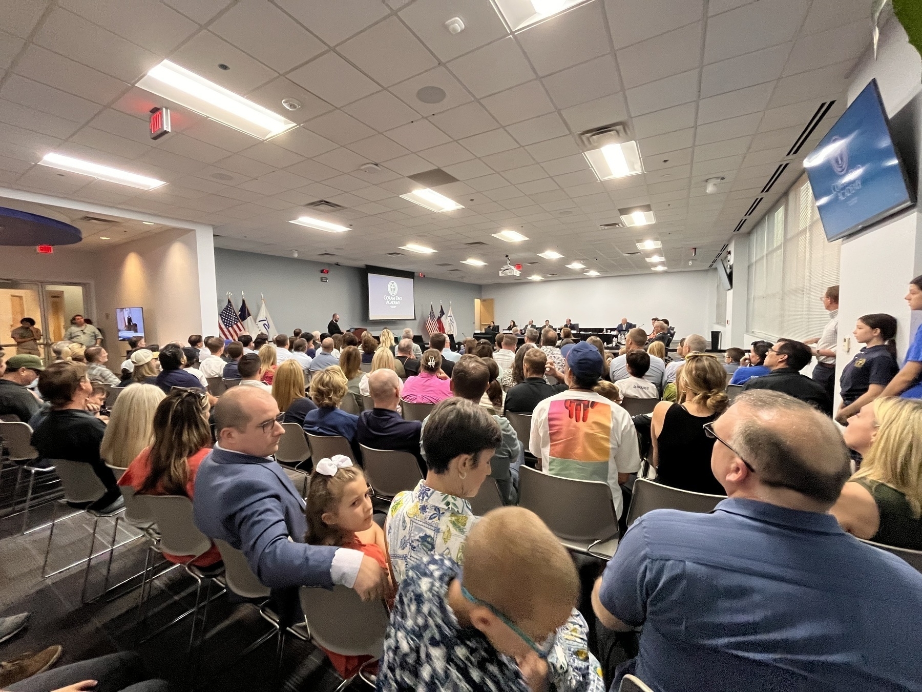 A large group of people is gathered in a conference room, attentively facing a presentation area at the front.