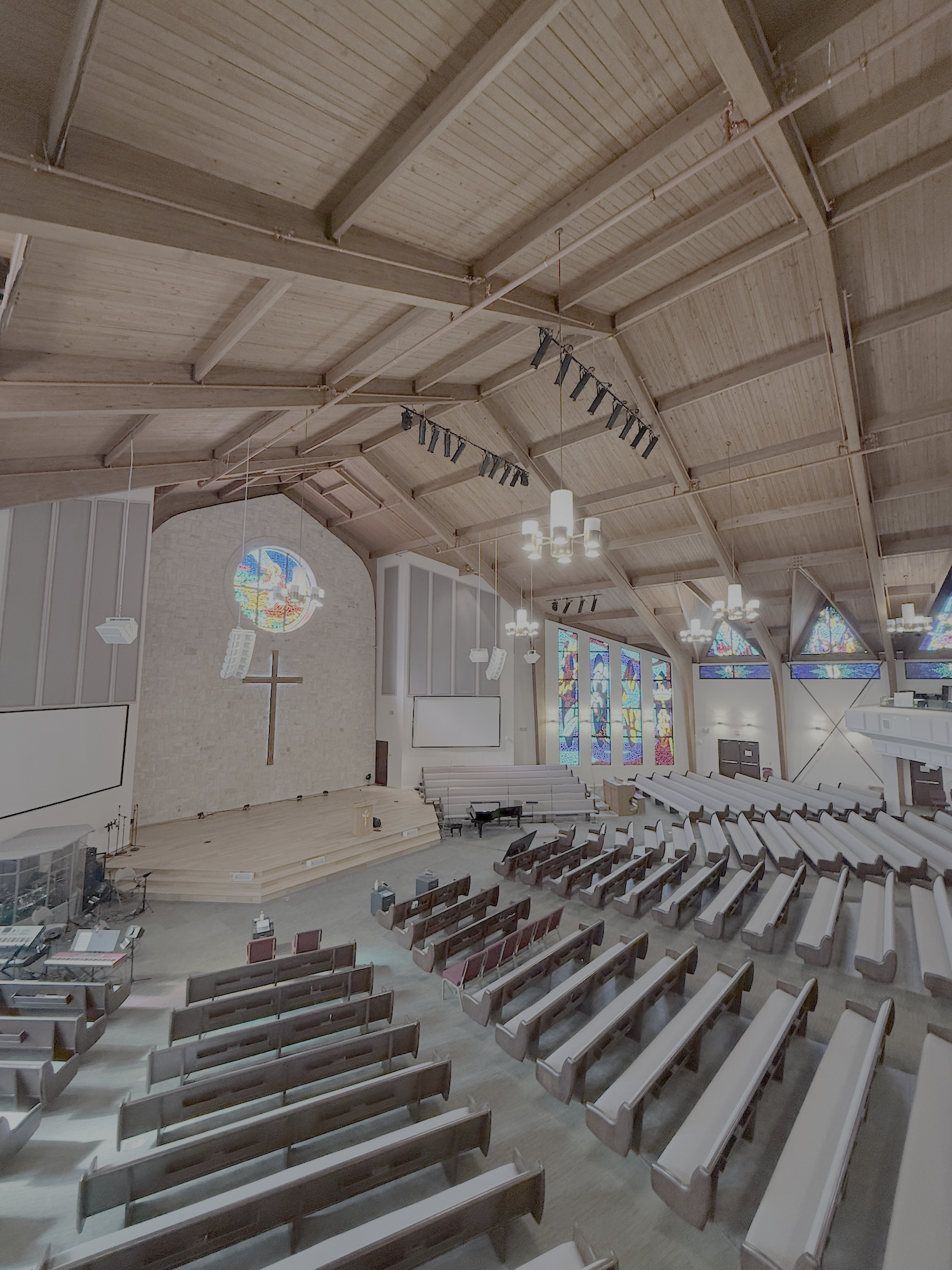 A spacious church interior features wooden beams, a large cross on the stone wall, stained glass windows, and rows of empty pews.