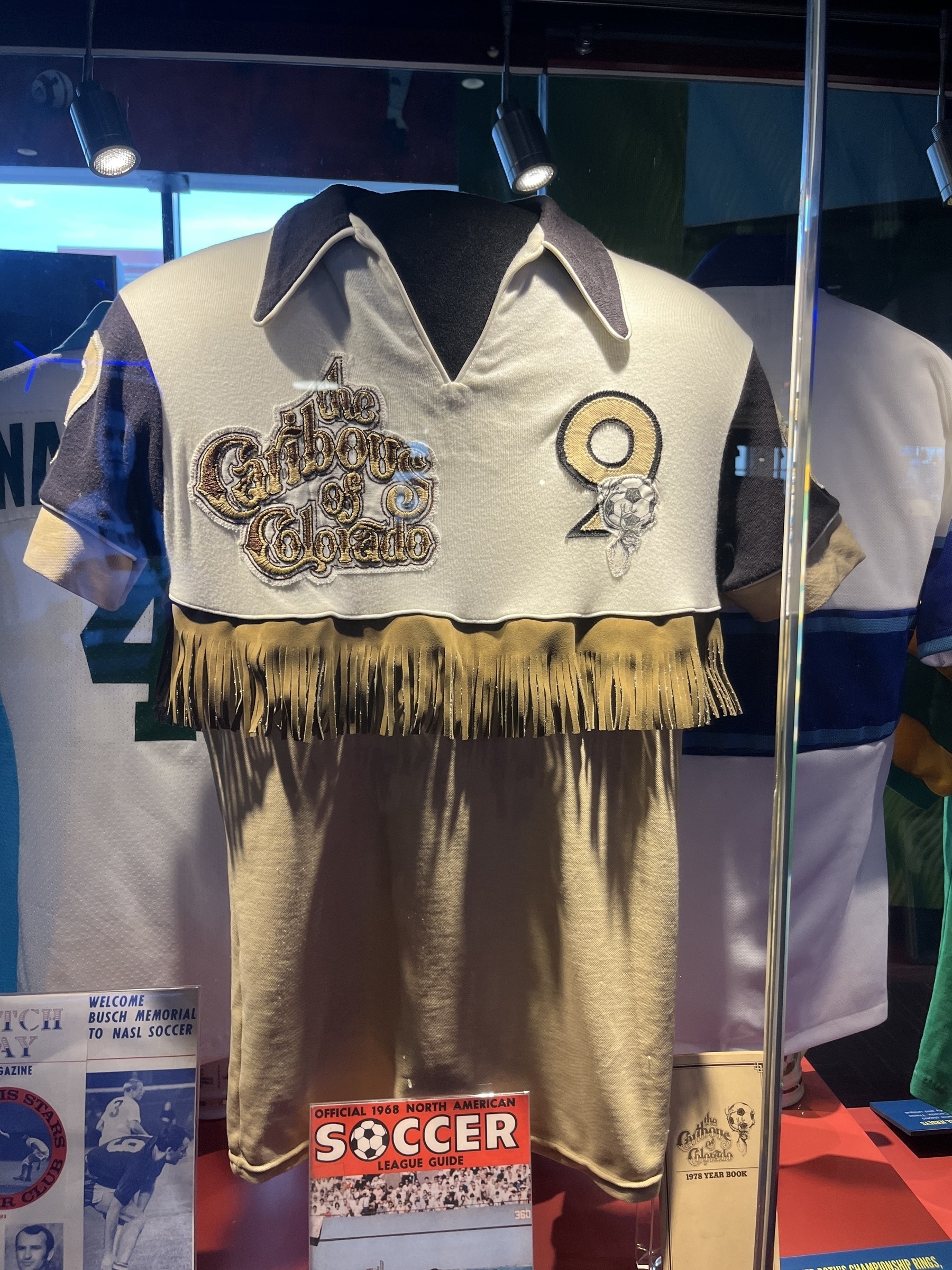 A vintage soccer jersey with fringe detailing and the text Caribous of Colorado displayed in a glass case.