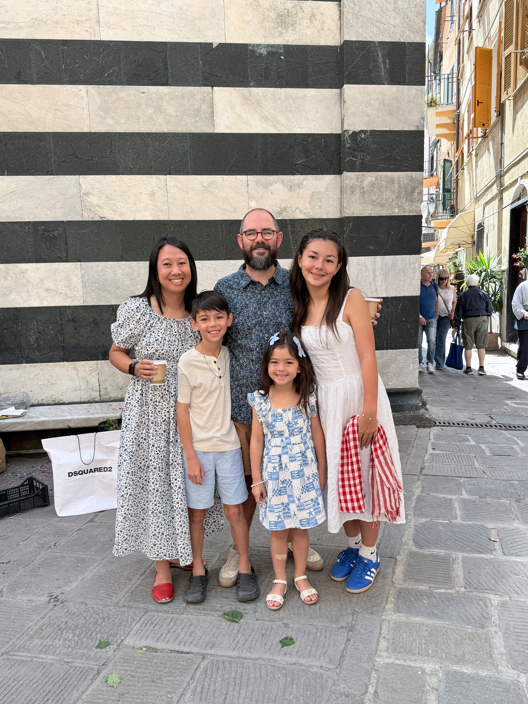 A family of five, including three children, is smiling and standing together in a cobblestone street, surrounded by buildings.